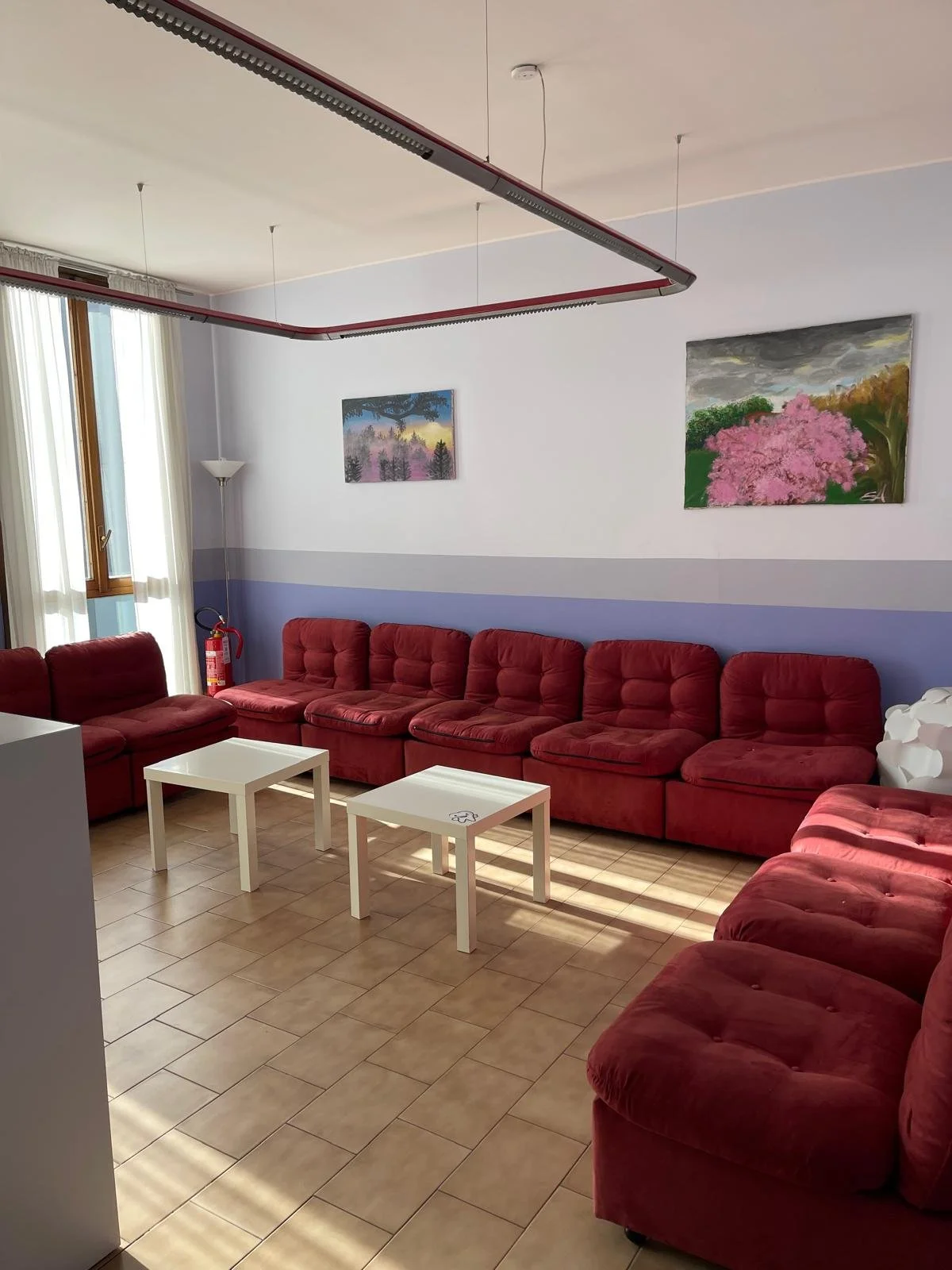 Sala di soggiorno con divani rossi, due tavolini bianchi, pareti bianche con una fascia azzurra, quadri floreali alle pareti, finestra con tende chiare, lampada da terra.