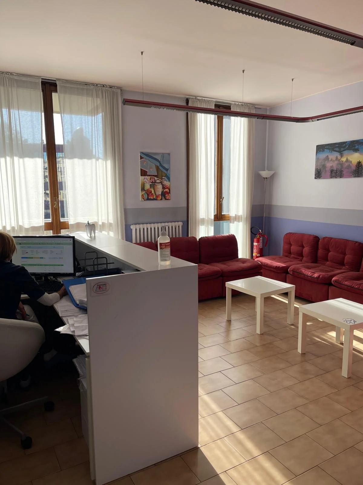 Sala d'attesa con divani rossi, due tavoli bianchi, grandi finestre con tende trasparenti, pareti dipinte di bianco e azzurro, quadri decorativi, lampada da terra, estintore.