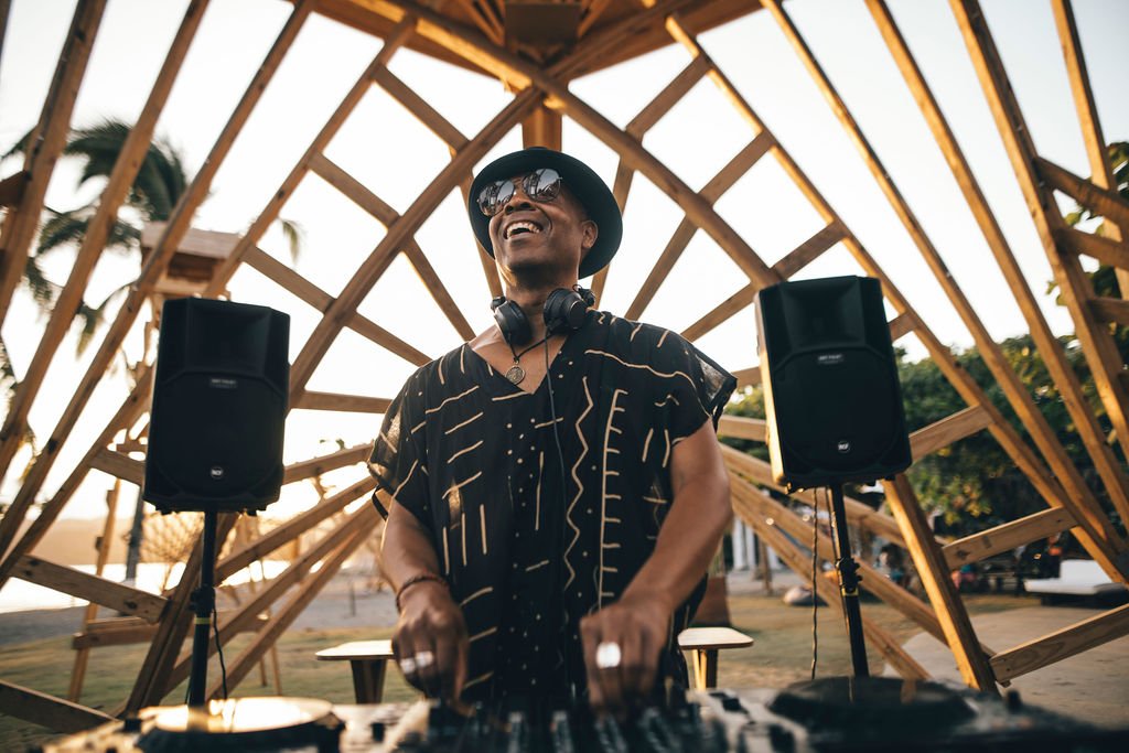 Marques Wyatt, wearing sunglasses, a hat, and headphones around his neck, smiling while performing outdoors at sunset with palm trees and a wooden structure in the background.