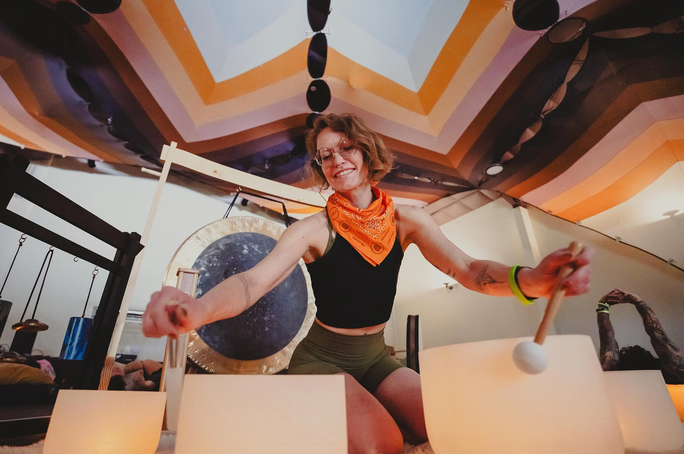 A woman with curly hair, glasses, and a bright orange bandana around her neck plays crystal bowls in a room with colorful, artistic ceiling and moon photo.