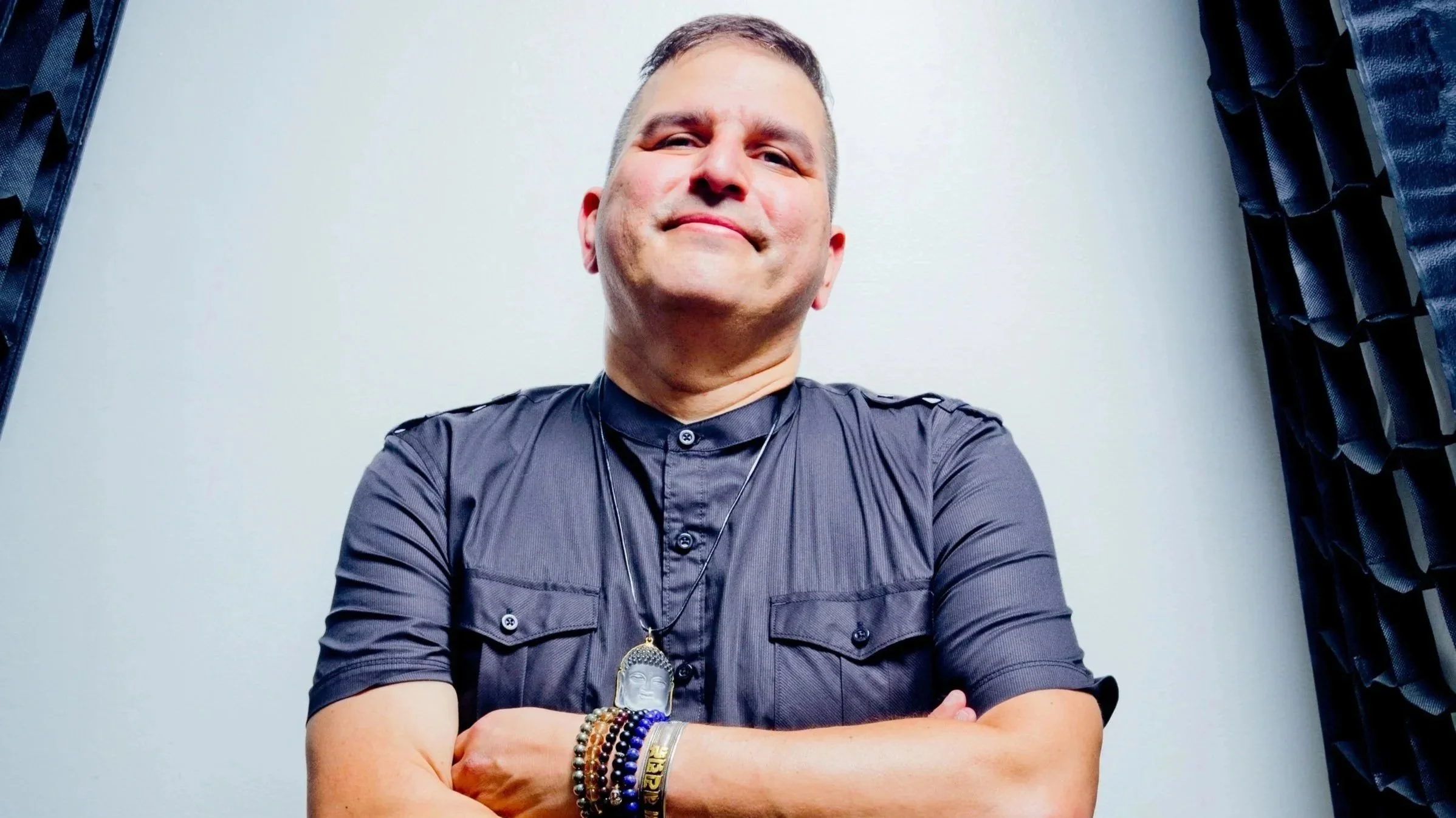 Doc Martin, wearing a dark shirt with rolled-up sleeves, standing with arms crossed, in front of a plain background flanked by dark curtains.