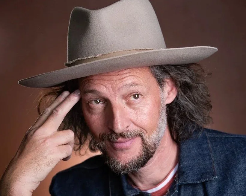 DJ Garth, wearing a gray wide-brimmed hat and a denim jacket, posing with his hand touching the side of his head against a brown background.