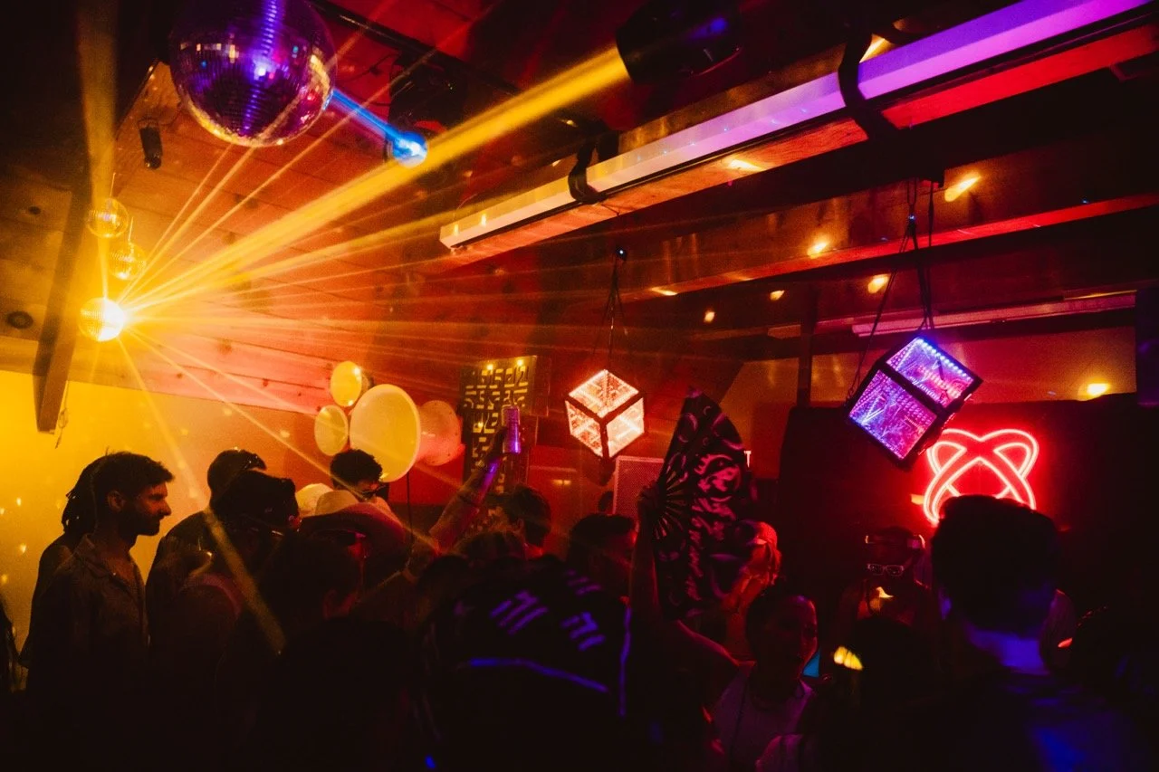 People dancing and socializing in a dimly lit nightclub with colorful lights, a disco ball, and hanging geometric decor.