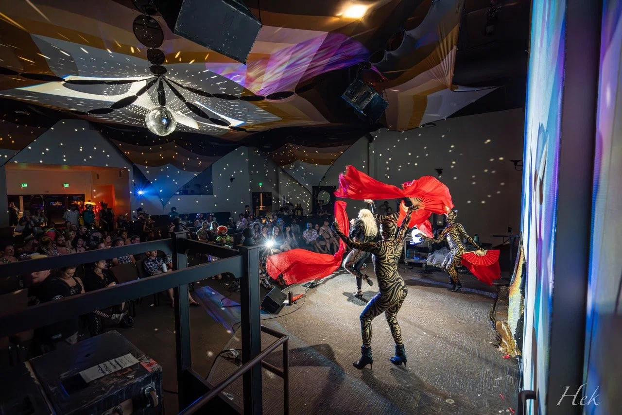 Performers with red fans and colorful costumes dancing on stage at a nightclub with an audience watching, disco ball overhead, and colorful lighting effects.
