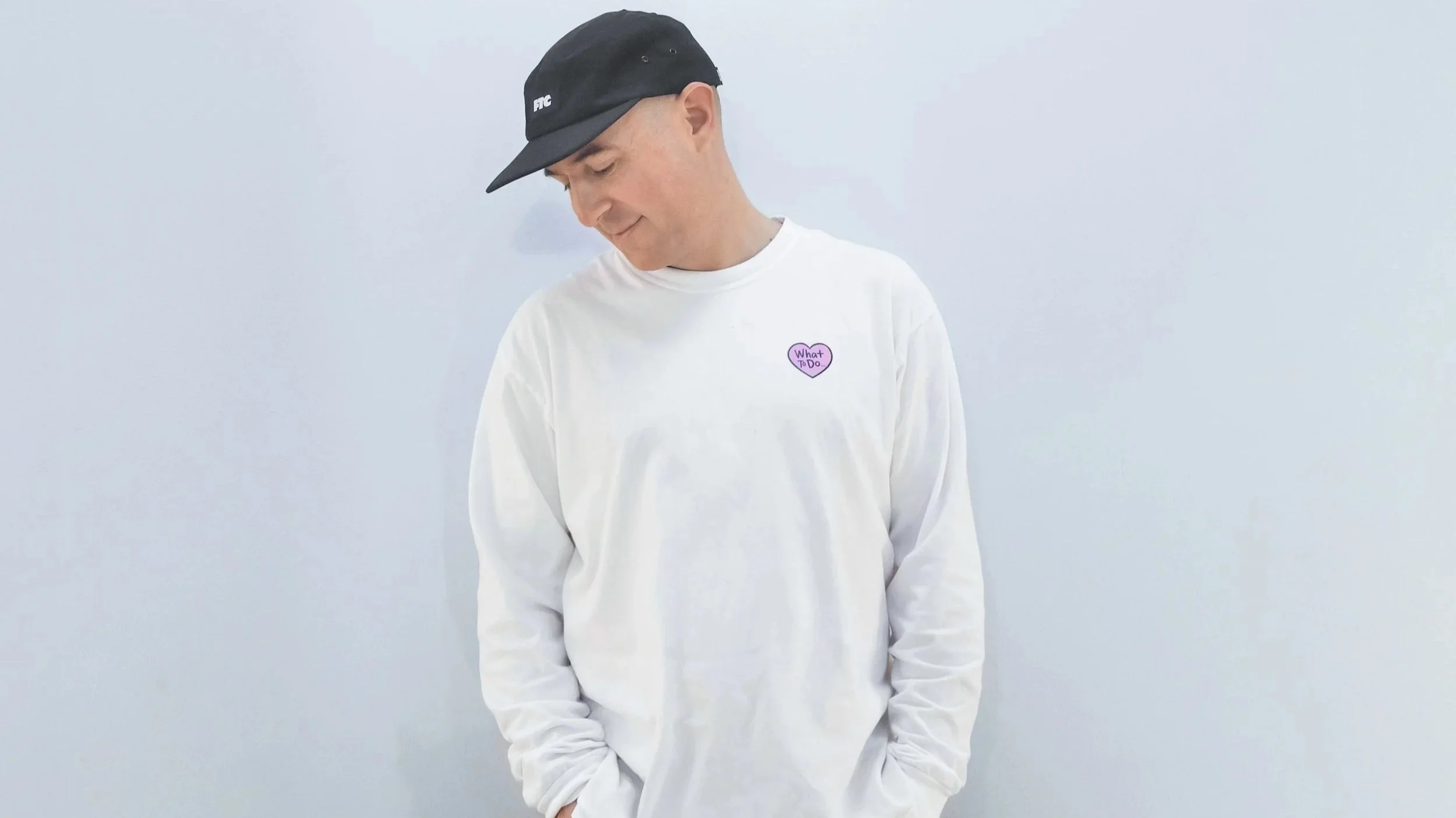 Justin Martin, wearing a black cap and a white sweatshirt with a purple heart-shaped pin that says 'What To Do'. He is looking down and smiling softly against a plain light-colored wall.