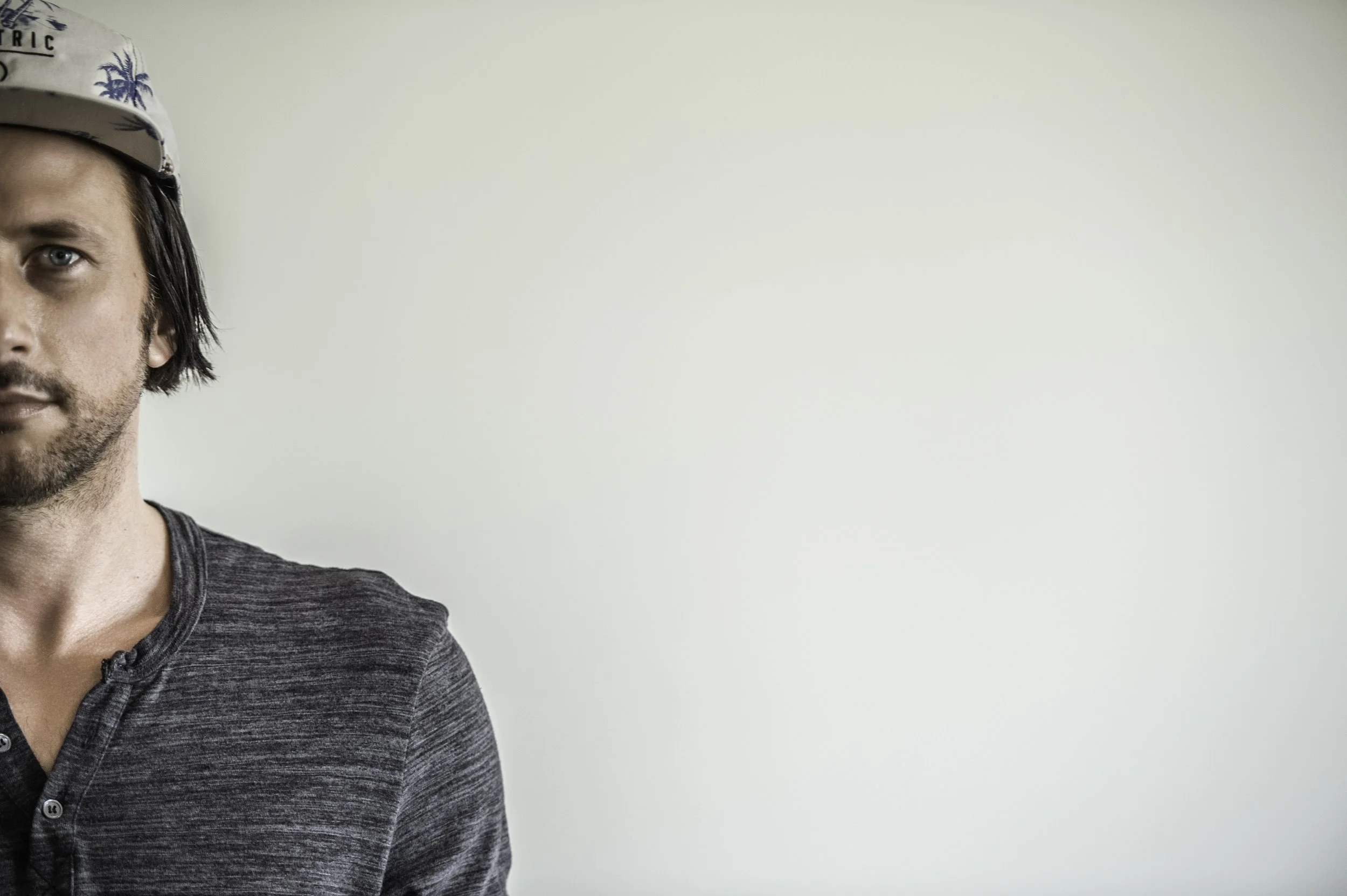Close-up of Miguel Migs, wearing a gray shirt and a cap with palm tree and surfboard pattern, standing against a plain light-colored wall.