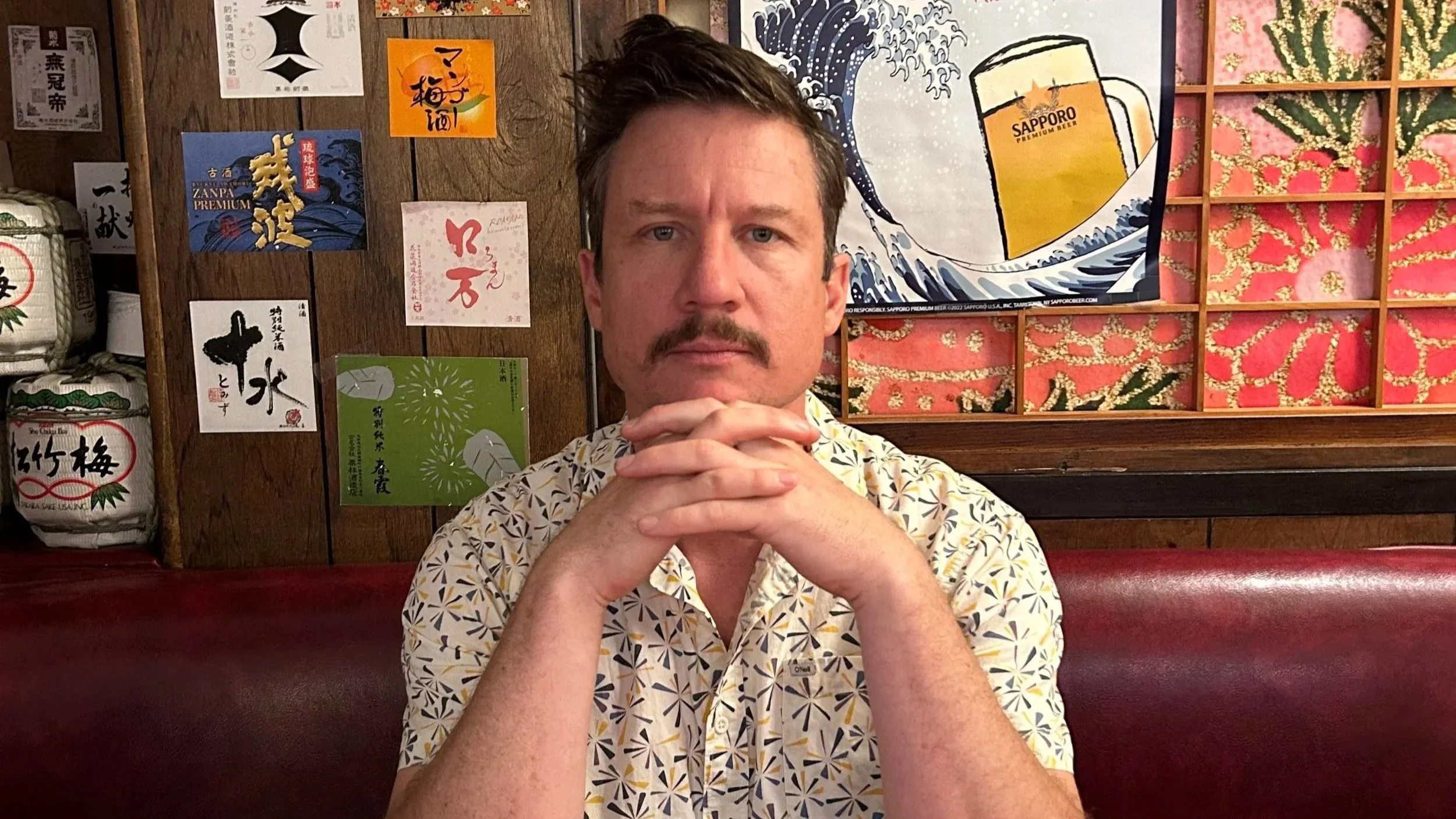 Om Records artist Rithma, sitting in a Japanese-themed restaurant. He is wearing a patterned short-sleeve shirt and resting his hands under his chin. The background features Japanese posters, a wave artwork, and a Sapporo beer poster.