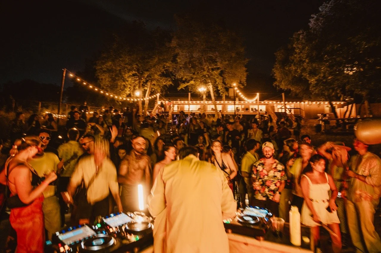 People dancing and partying outdoors at night with string lights and trees, with a DJ playing music in the foreground.