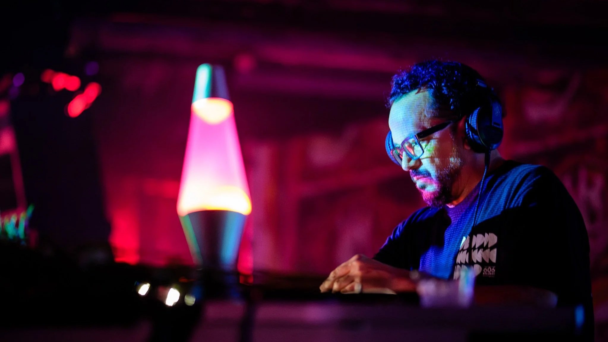 DJ Mark Farina (Mushroom Jazz), focused while operating DJ equipment in a dimly lit room with colorful lighting and a lava lamp in the background.
