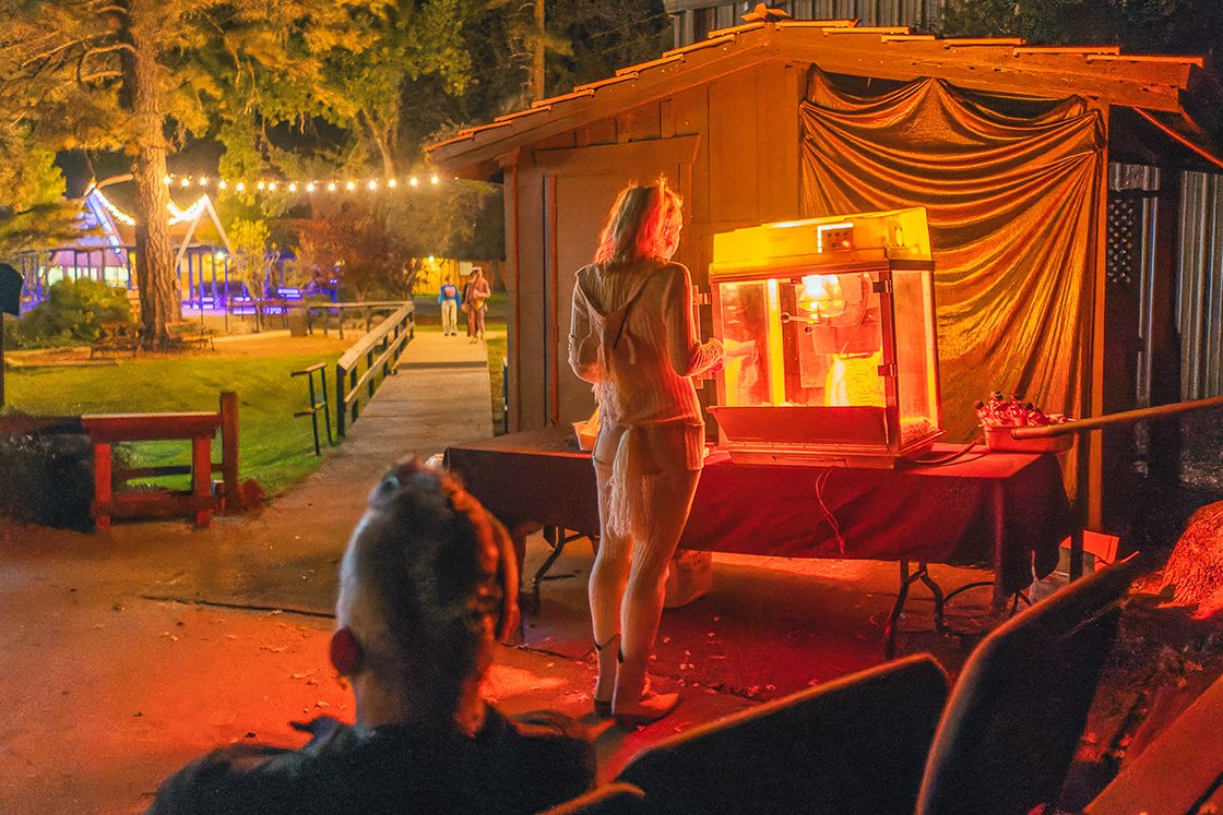 A woman stands at an outdoor carnival game booth at night, illuminated by the red glow of a ticket dispenser, with a dog sitting nearby in foreground, and a playground with string lights in the background.