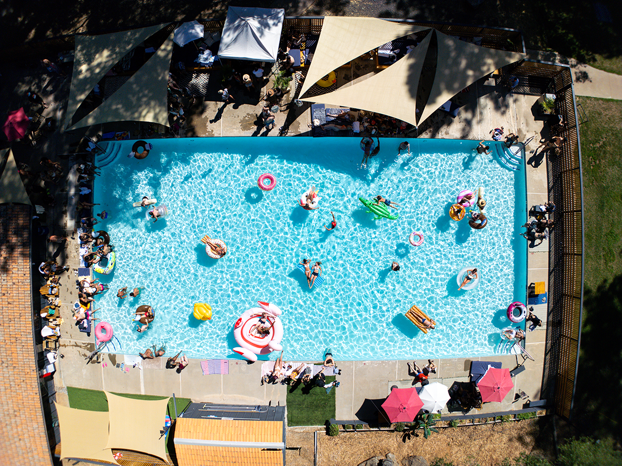 Aerial view of a crowded swimming pool with people swimming, floating on inflatables, and relaxing on pool floats, surrounded by umbrellas and chairs.