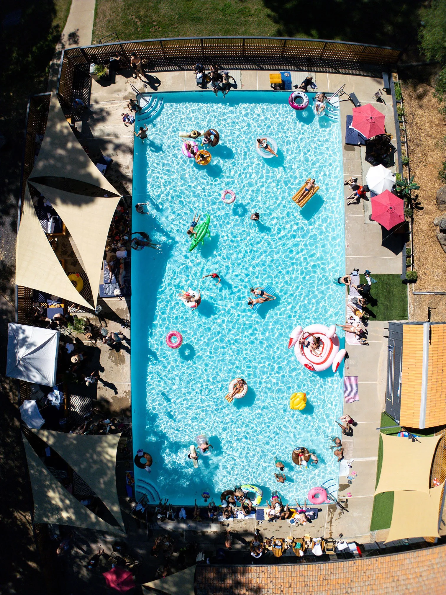 A crowded swimming pool filled with people, floats, and pool toys. Some people are swimming, floating on inflatables, or sitting at the edge. The pool is surrounded by shaded areas, umbrellas, and poolside furniture.