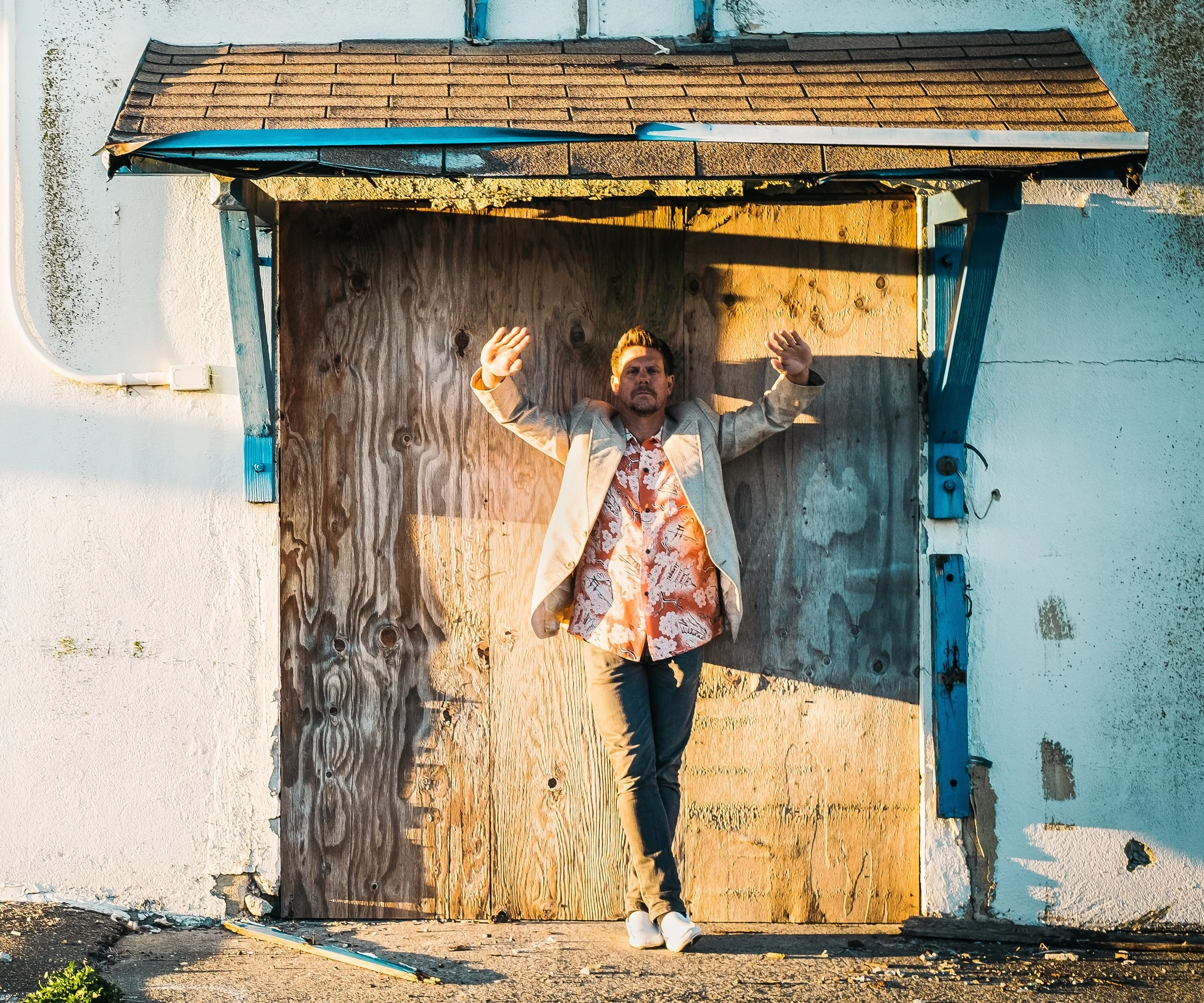 Shiny Objects standing with arms raised in front of a wooden boarded-up entrance, with sunlight illuminating his face and clothes.