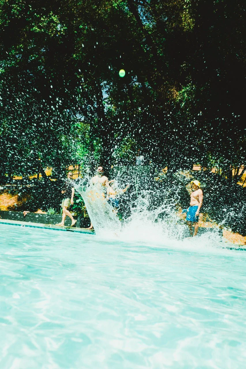 People playing and splashing in a swimming pool on a sunny day, with trees in the background.