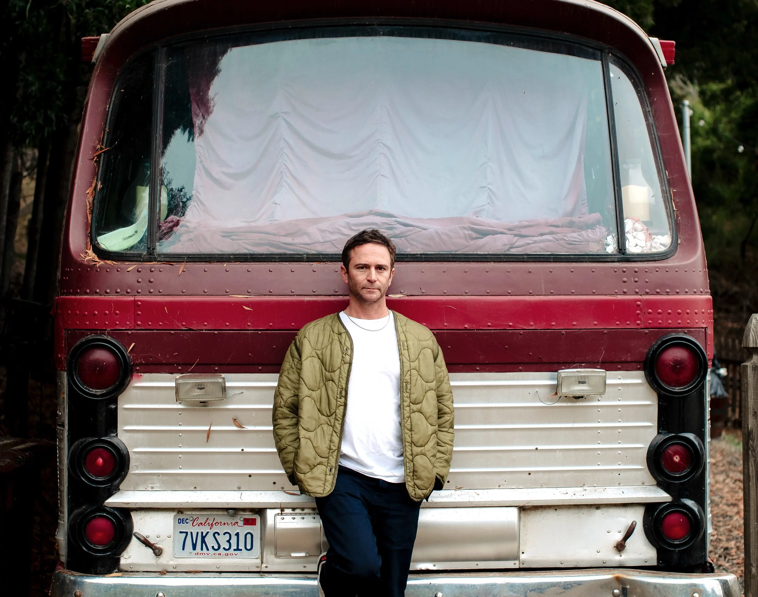 Galen (Sunset Soundsystem) stands in front of a vintage red and white bus with a California license plate, outdoors among trees.