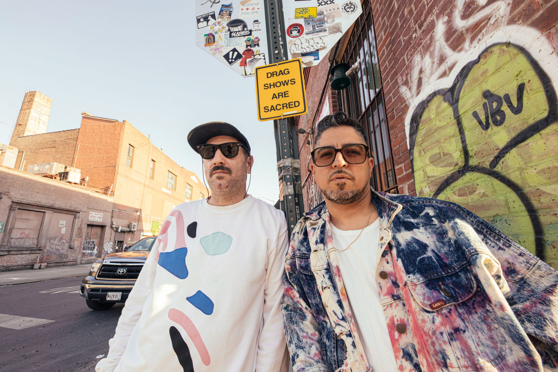 Pillowtalk, in front of graffiti on a brick wall and a street scene with cars and buildings.