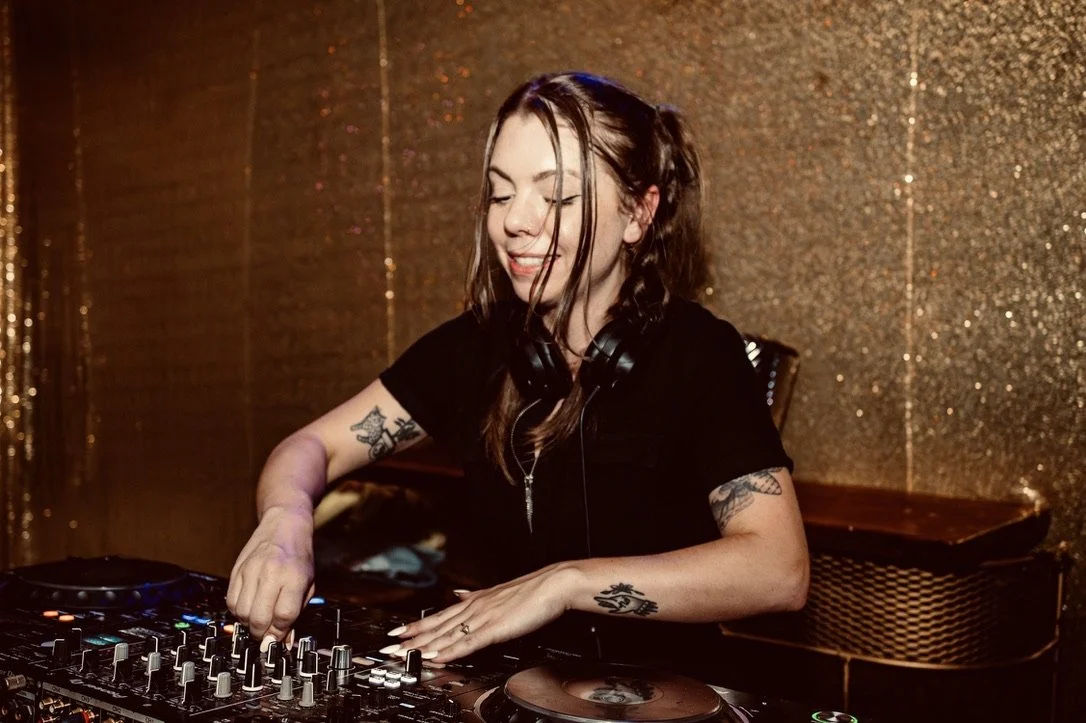 A young woman with tattoos and long hair, wearing headphones, is DJing at a party with a golden, glittery wall backdrop.