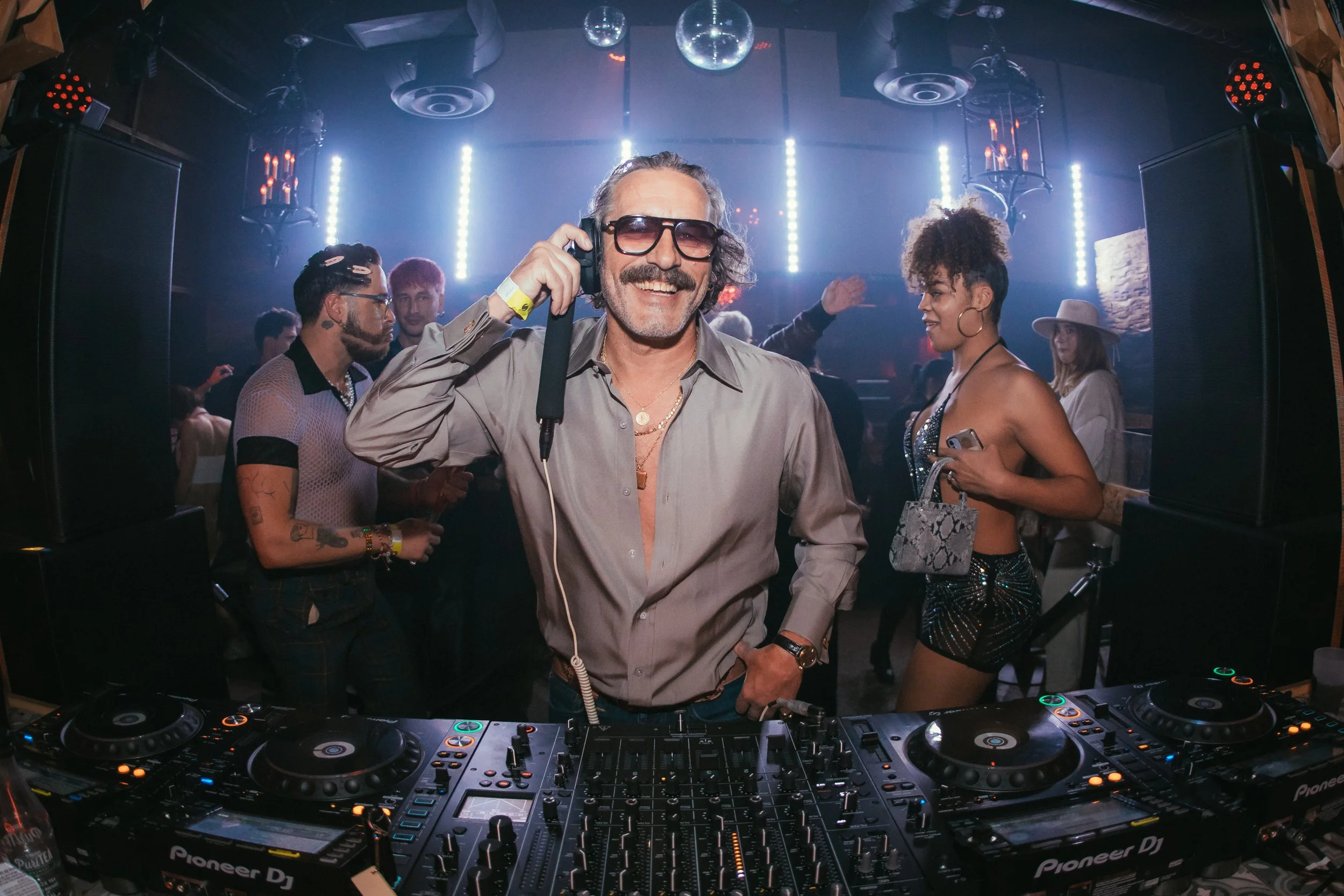 DJ Harvey in sunglasses and gray shirt smiling and talking on headset at a nightclub with people dancing and socializing around him, colorful lights in the background.