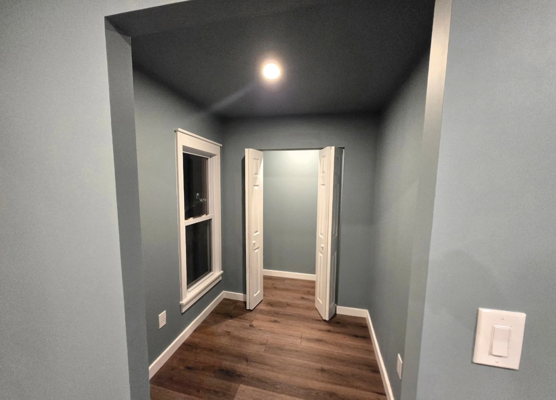 Empty room with light blue walls, a window, and white double doors leading to a small closet or passage. Wooden floor and a ceiling light.