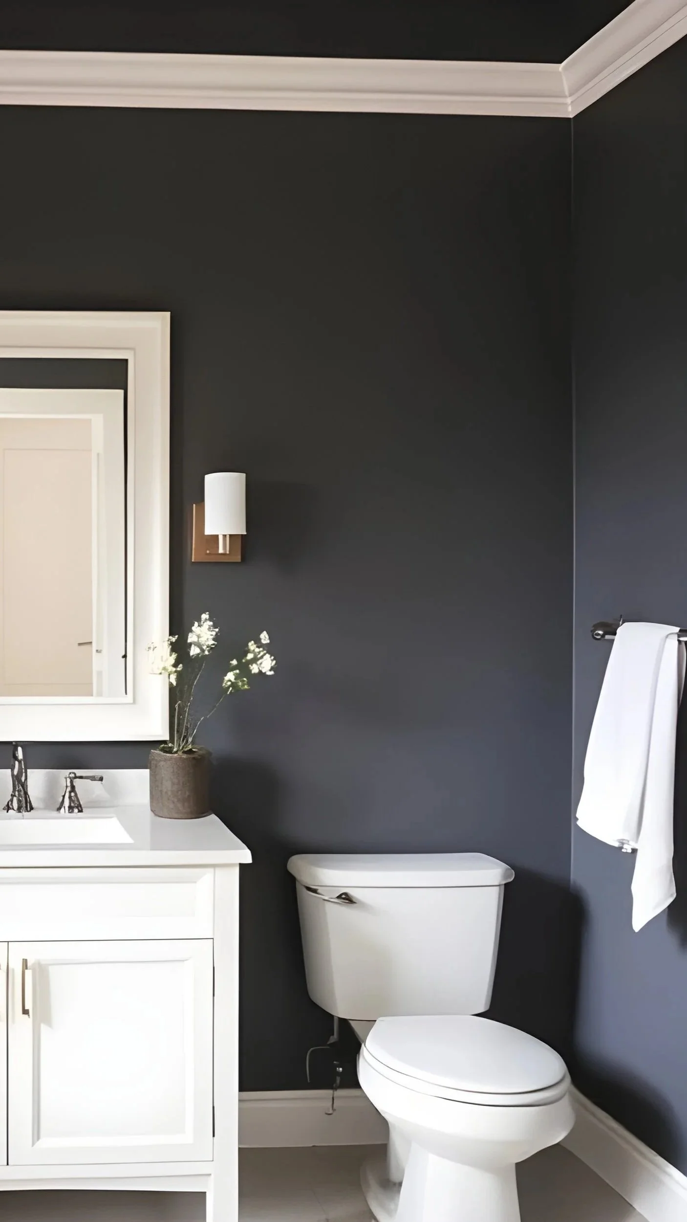 Modern bathroom with dark gray walls, white vanity with black fixtures, a rectangular mirror, a wall-mounted light fixture, a white toilet, a towel hanging on a rod, and a decorative bowl with flowers on the vanity.