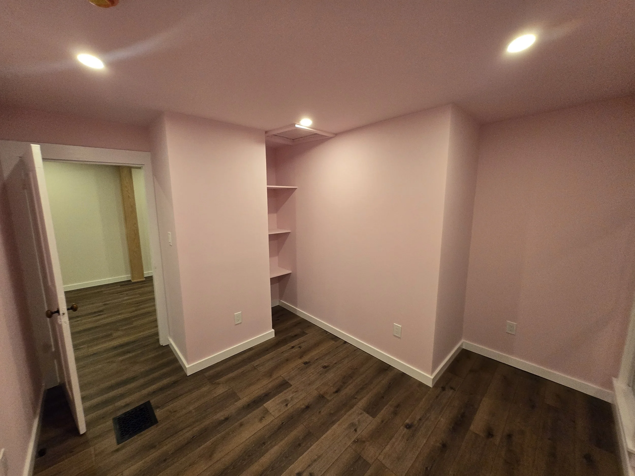 Empty pink-walled room with wooden floor, built-in shelves, and door leading to another room.