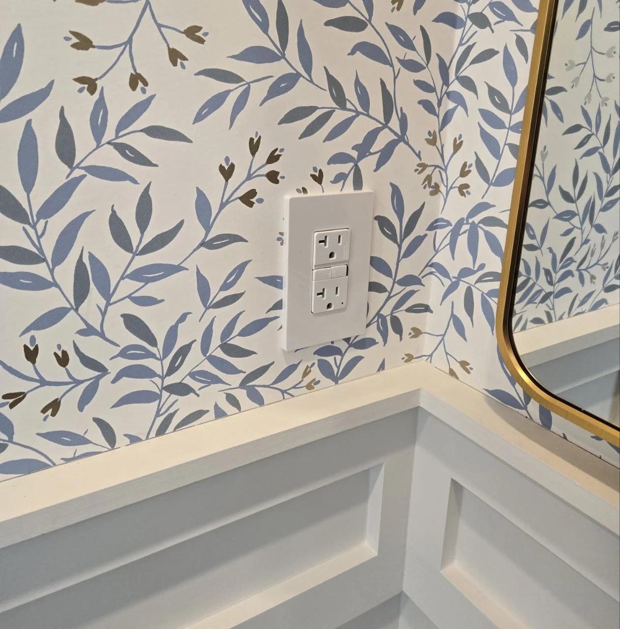 A close-up of a wall with blue and brown leaf-patterned wallpaper, an electrical outlet with two sockets, and a mirror with a gold trim.
