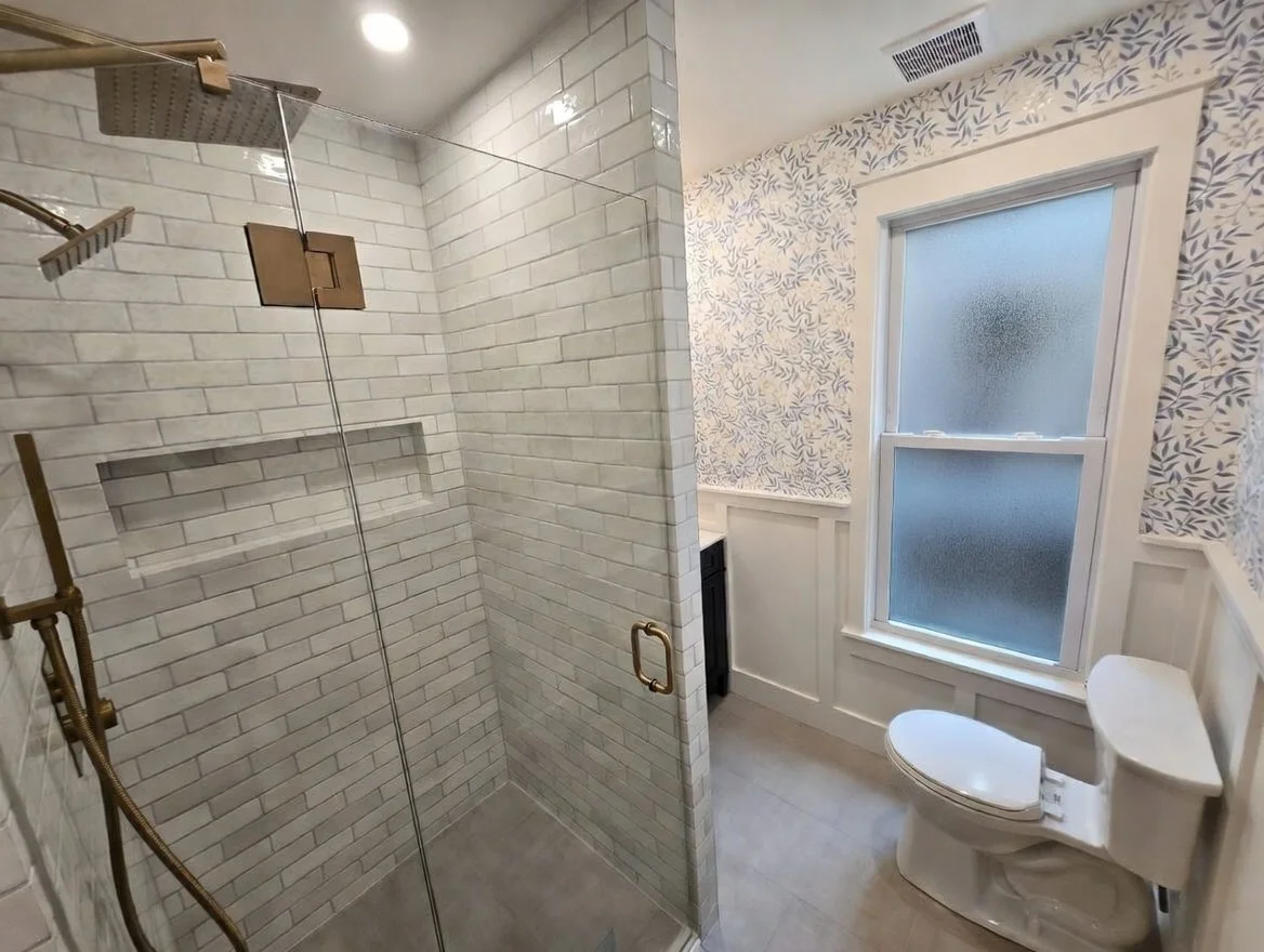 Bathroom with a glass-enclosed shower featuring white subway tile walls, a rain showerhead, a handheld shower, and a built-in niche; adjacent is a toilet with a frosted window and wallpaper with a subtle leaf pattern.
