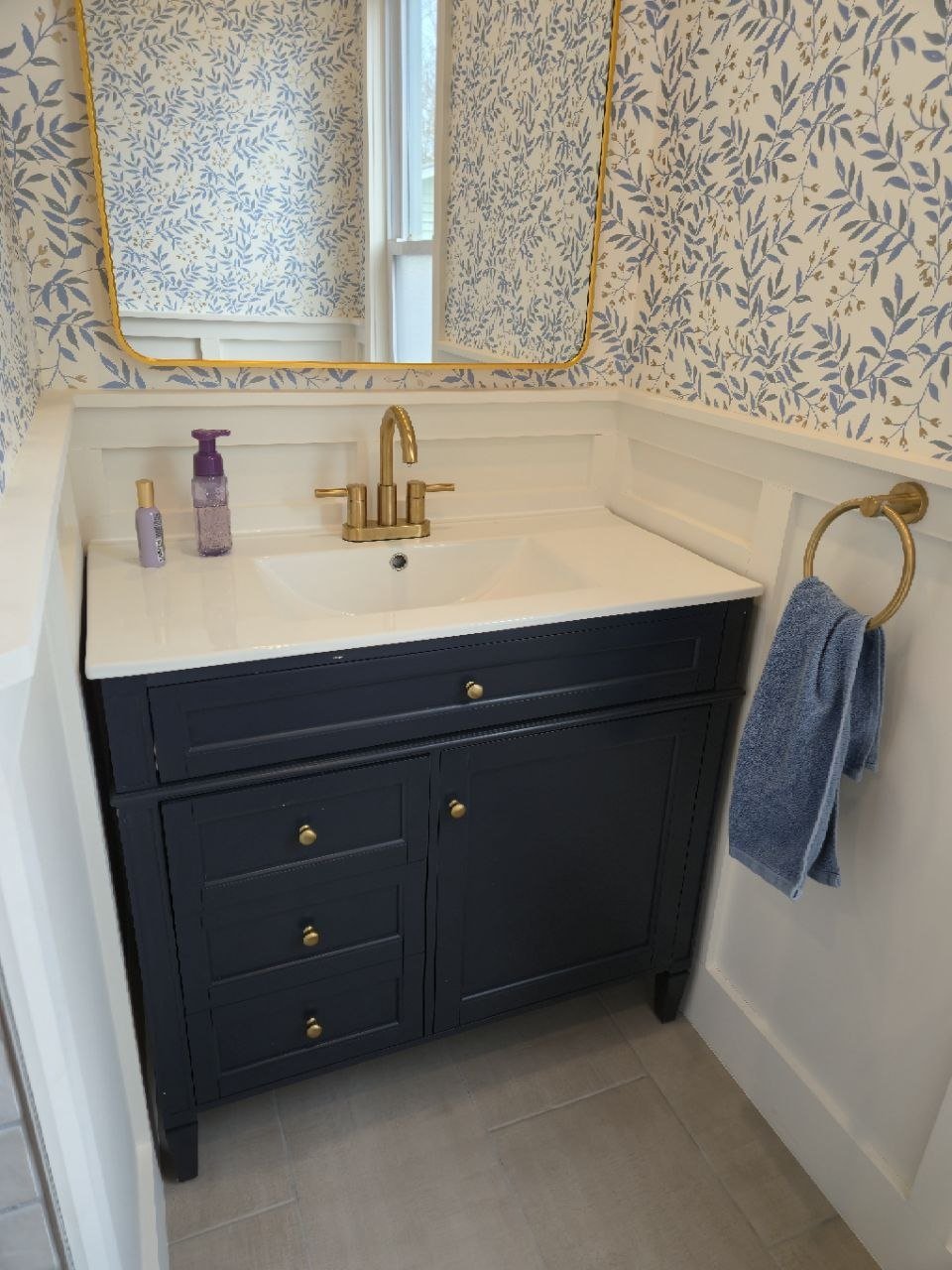 Bathroom vanity with a navy blue cabinet, a white countertop and sink, a gold faucet, a mirror with a gold frame, two bottles on the countertop, and a blue towel hanging on a gold towel ring.