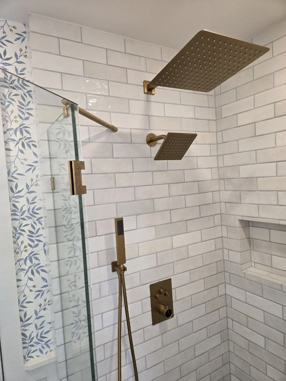 Bathroom shower with white subway tile walls, two gold square rain shower heads, a wall-mounted handheld shower with a flexible hose, and a built-in niche in the wall.