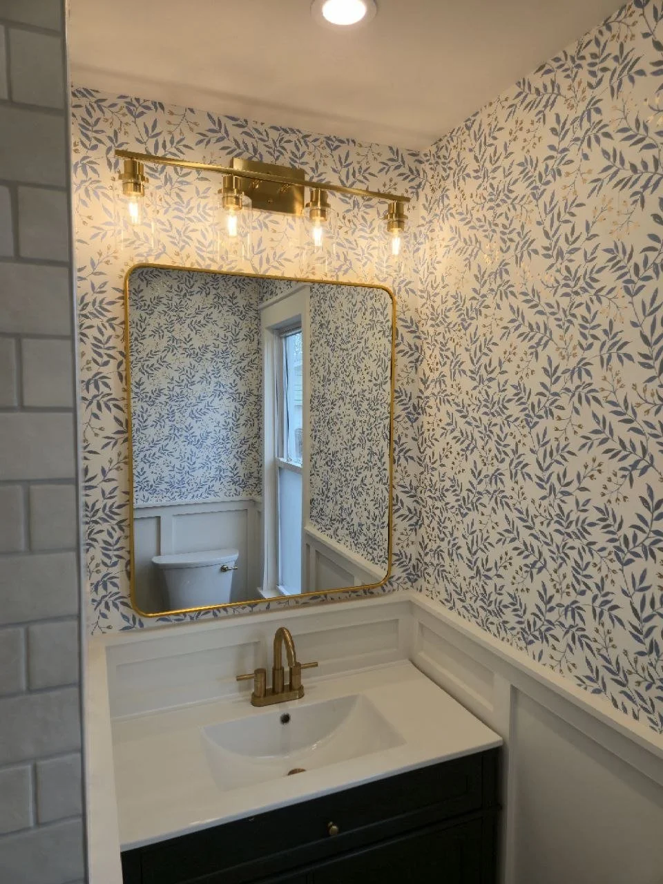 Bathroom with a decorative leafy wallpaper, a mirror with a gold frame, a white sink with gold fixtures, a black vanity, and a window reflected in the mirror.