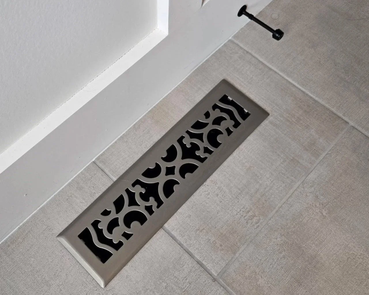 A metal floor vent cover with decorative cut-out patterns, installed on a tiled floor near the corner of a wall. There is a black nail or screw set into the wall beside it.