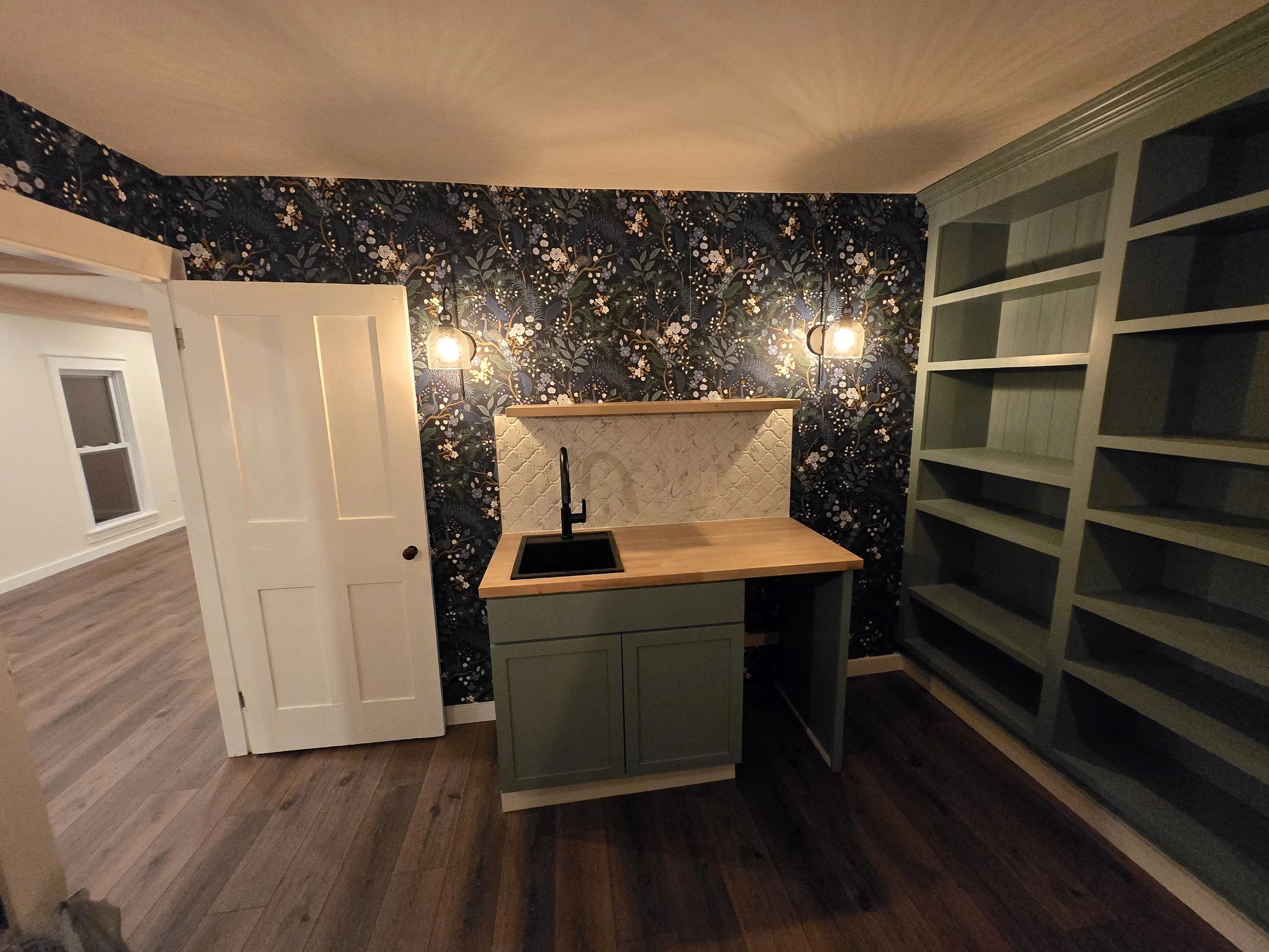 A small kitchenette with a black sink, green cabinets, and a wooden countertop, against a floral wallpapered wall with two wall sconces illuminating the space.