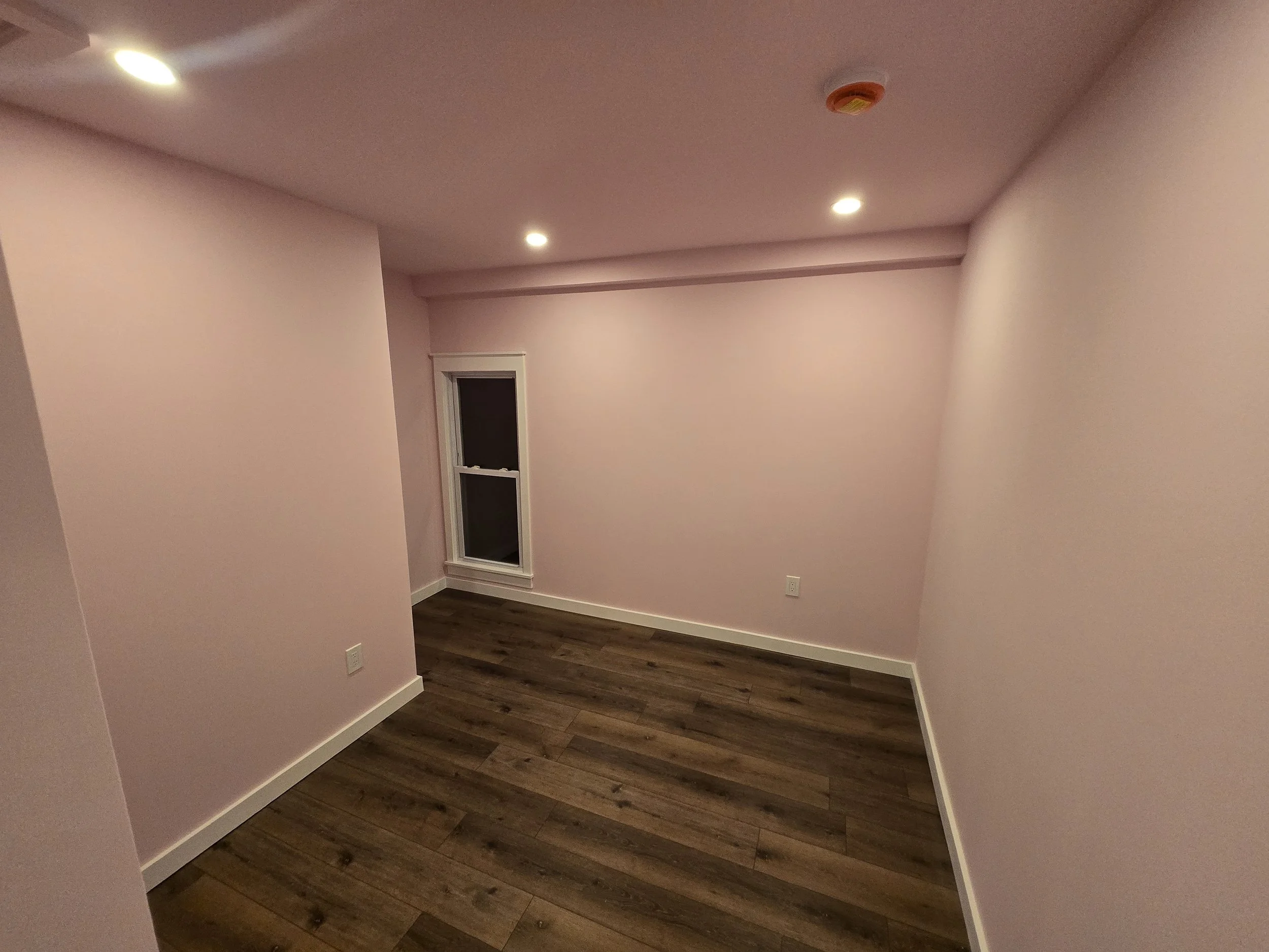 Empty room with pink walls, dark hardwood floor, small window, ceiling lights, and smoke detector.