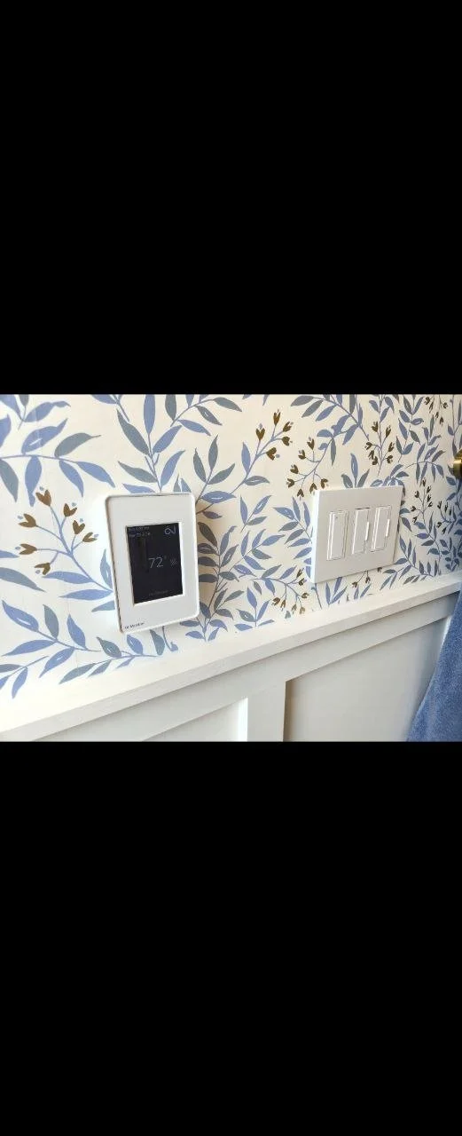 Close-up of a wall with floral wallpaper, a white digital thermostat displaying 72°F, and a white double light switch plate.
