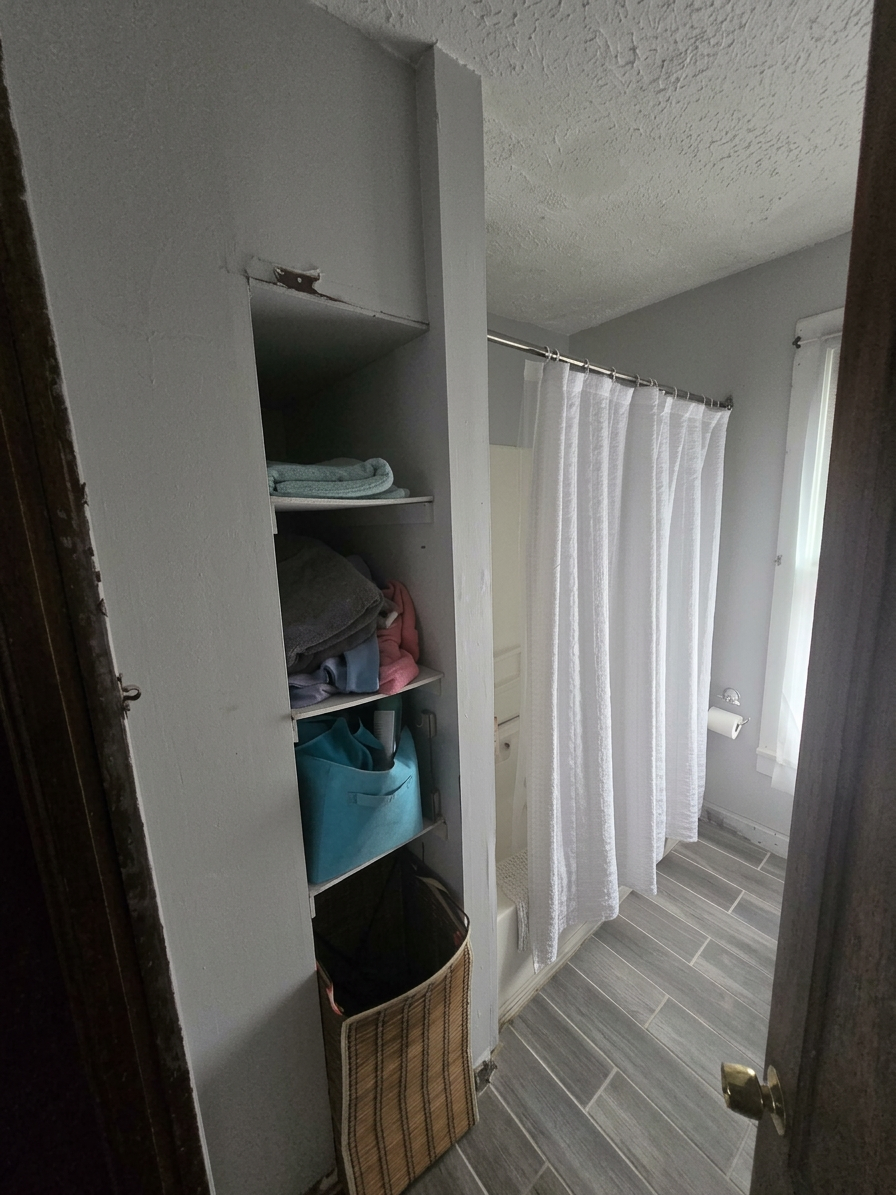 Small bathroom with gray walls, white shower curtain, and tiled floor, seen through an open wooden door. Shelves near the entrance hold folded towels and storage containers, with a laundry basket at the bottom.