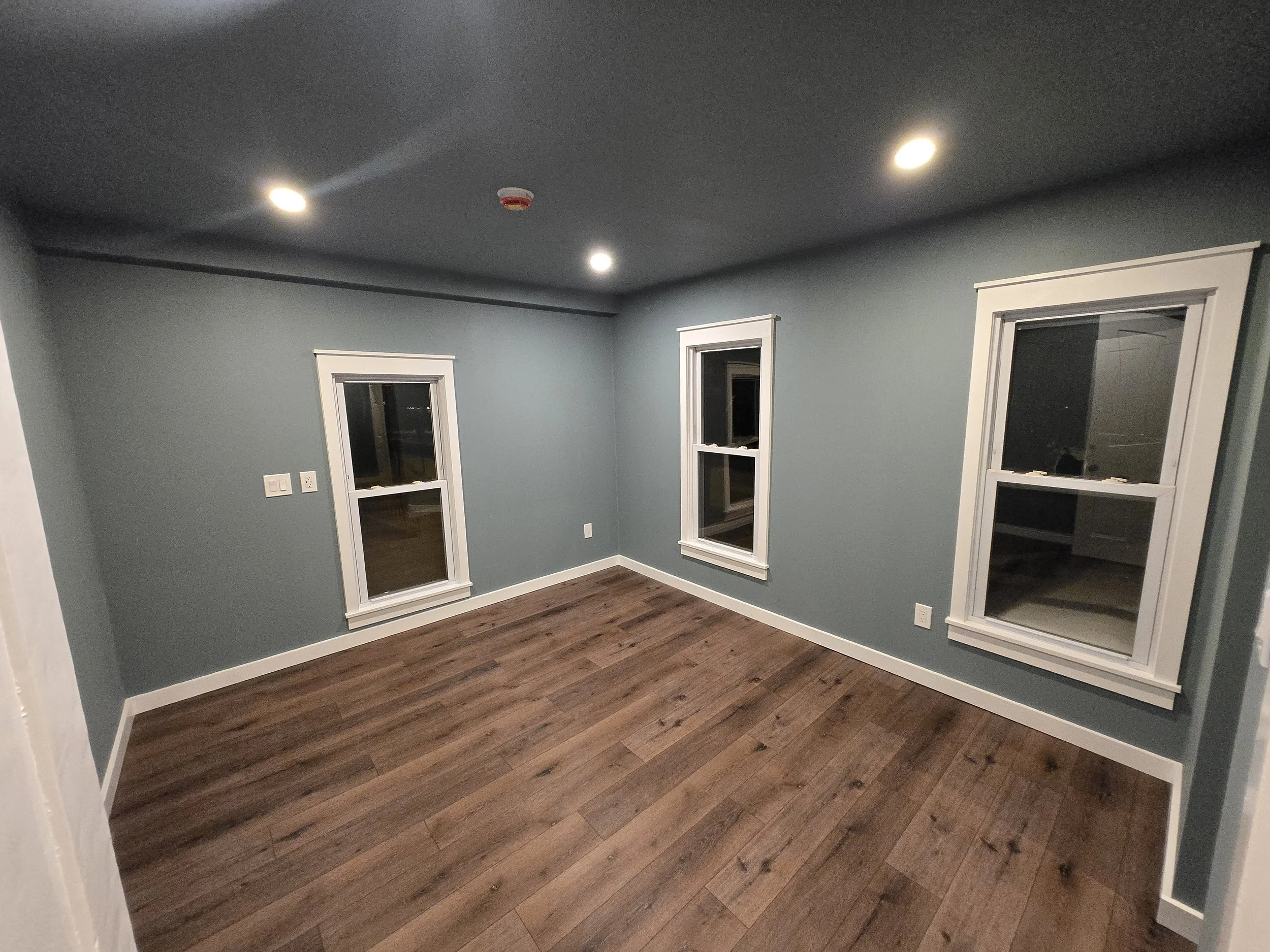 Empty room with three windows, hardwood floors, blue walls, and ceiling lights at night.