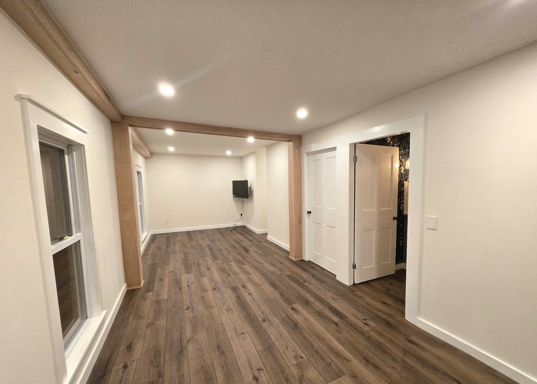 Empty living room with hardwood floors, white walls, ceiling lights, and windows, with a small wall-mounted TV in the back corner and an open doorway on the right.