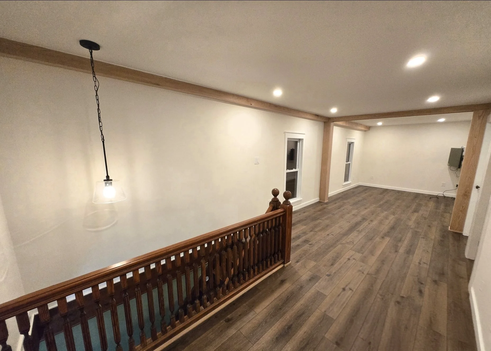 Empty living space with wooden flooring, white walls, recessed lighting, and large windows, with a wooden staircase railing visible.