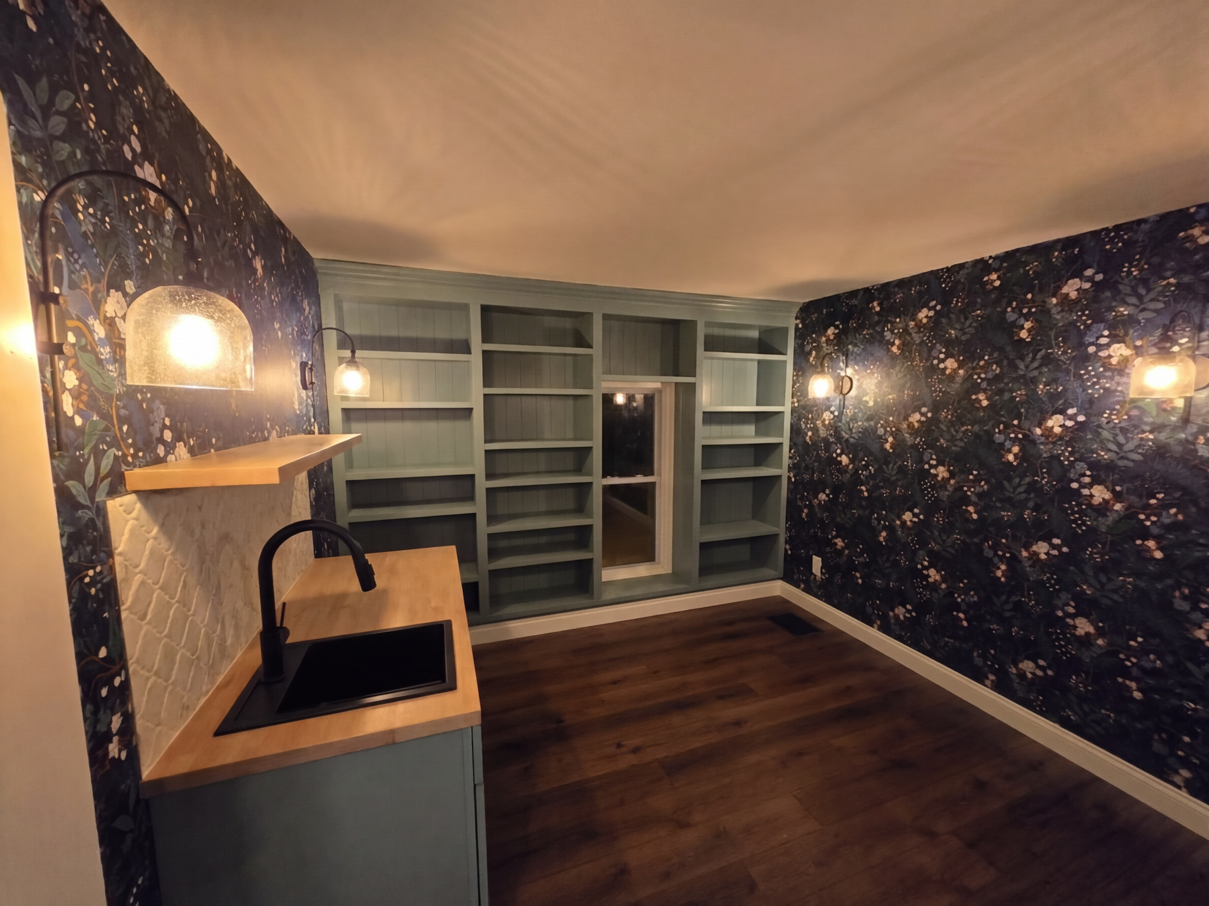 Empty room with dark floral wallpaper, built-in bookshelf, small wooden kitchen counter with a black sink, black faucet, and hanging wall lights.