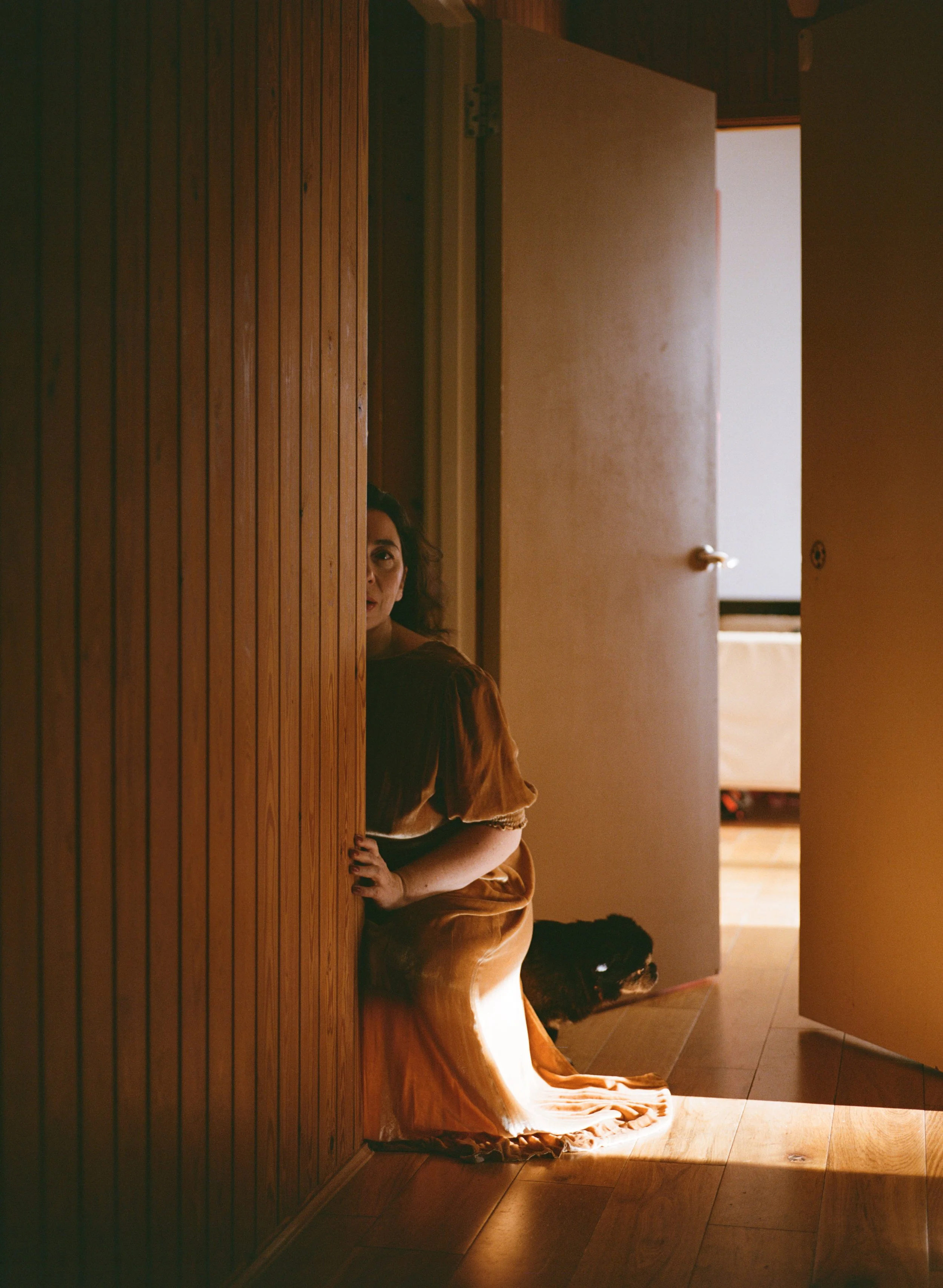 Woman with dark hair in an orange dress peeks out from behind a wooden wall in a dimly lit room, with a door slightly open revealing light outside.