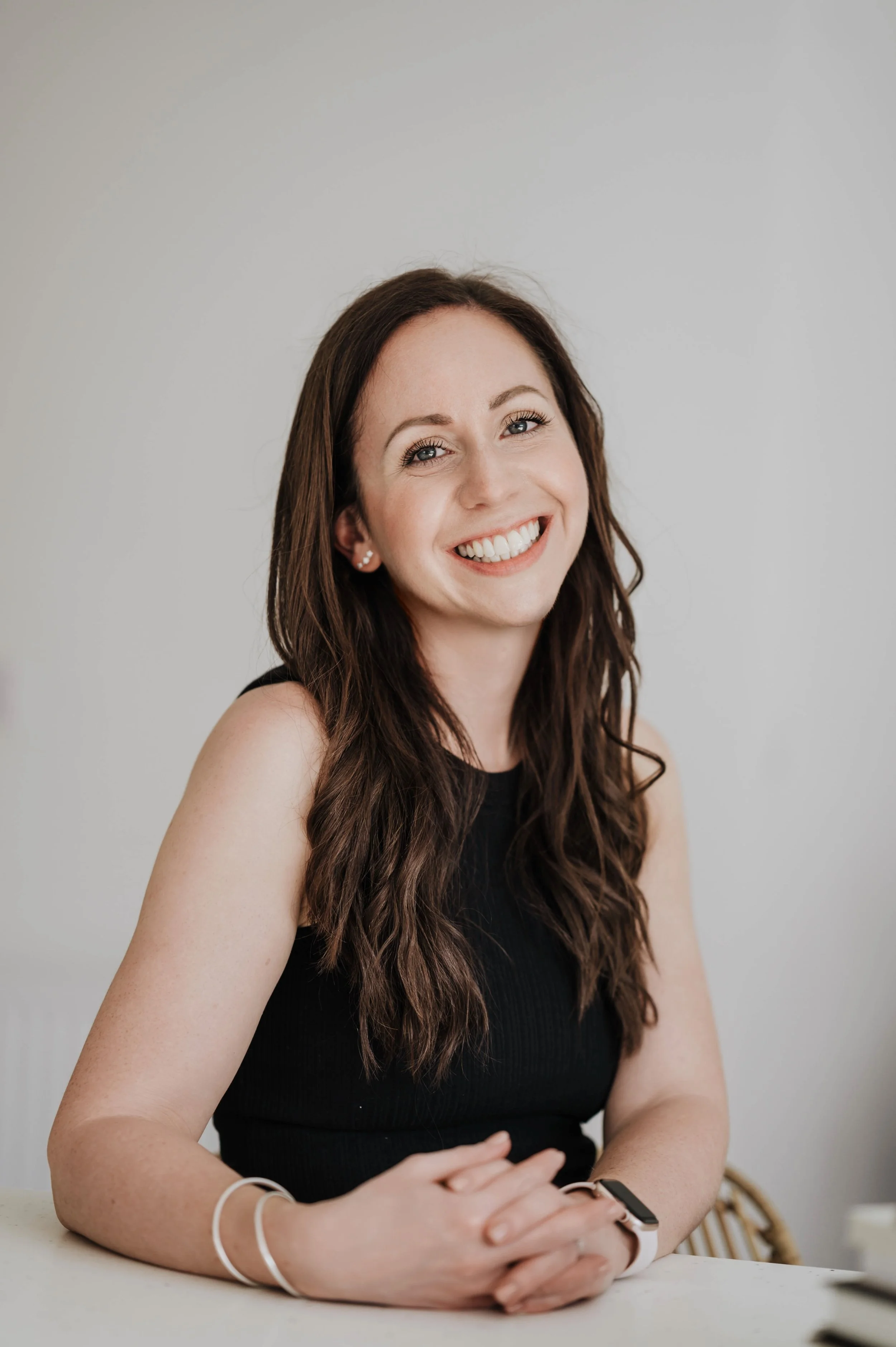 A young woman with long brown hair, wearing a black sleeveless top, smiling at the camera in a room with a neutral wall background.