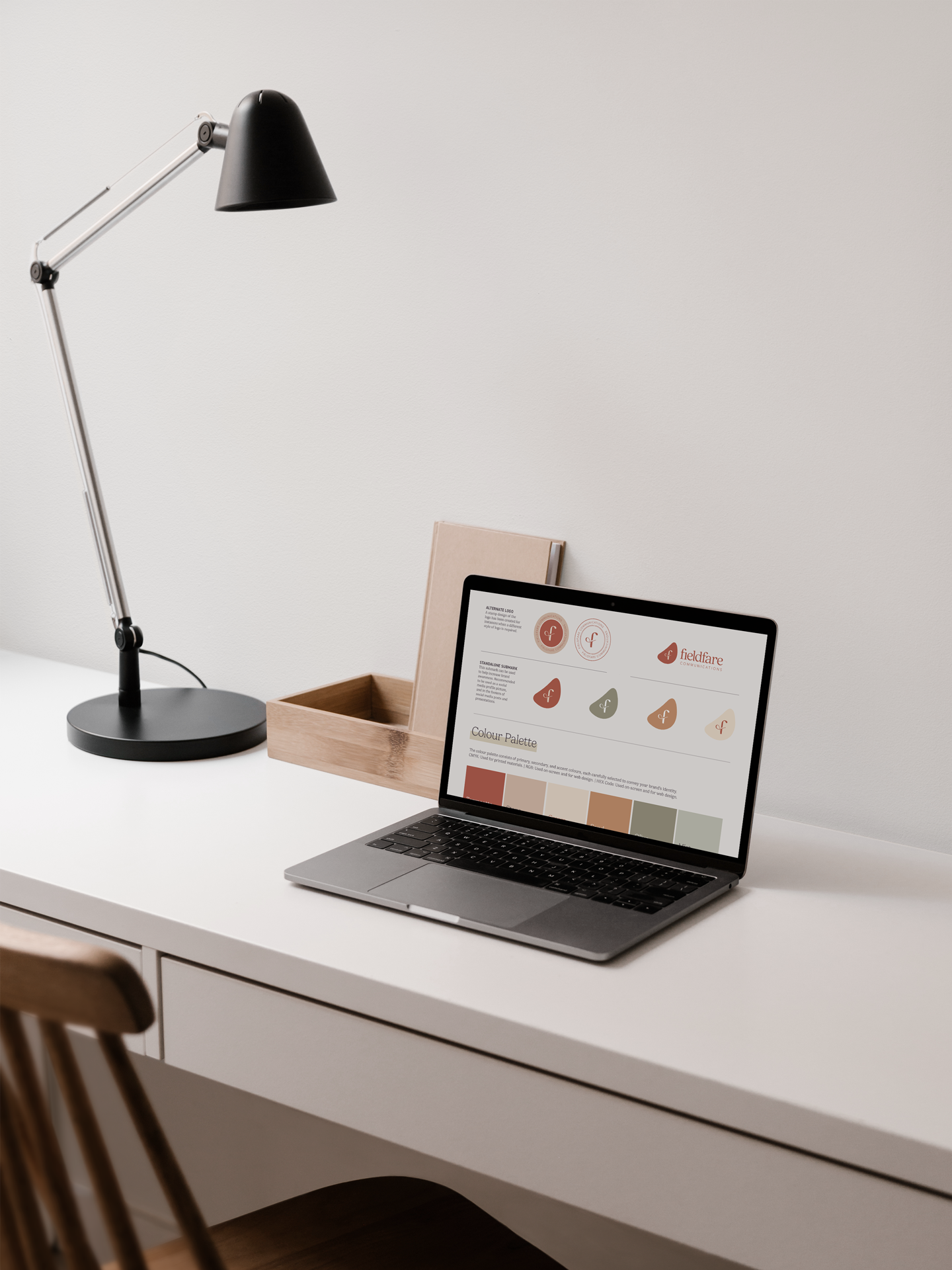 White desk with a silver laptop, a black desk lamp, a beige notebook, and a wooden tray holding a tablet displaying a brand color palette.