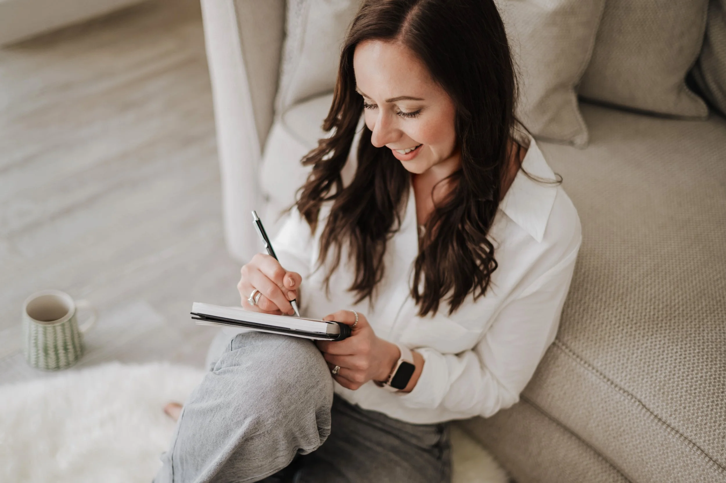 A woman with long brown hair sitting on the floor next to a light grey sofa, smiling while illustrating in a sketchbook with a pencil.