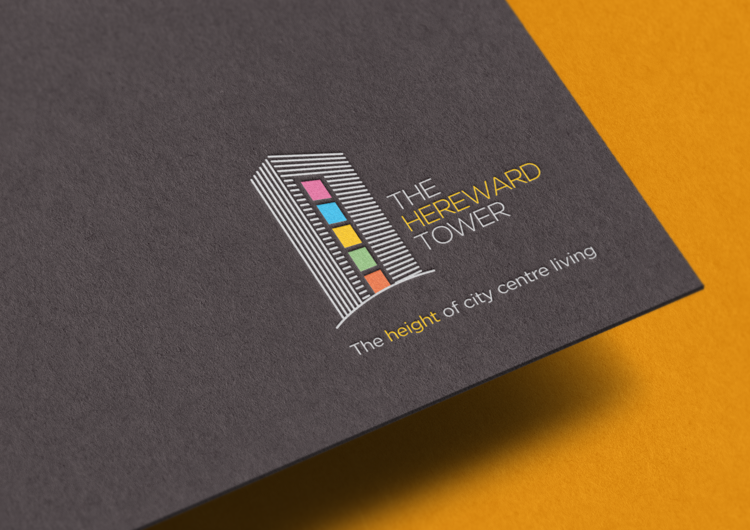 Close-up of a dark grey printed material with a colourful logo titled "THE HEREWARD TOWER" featuring a stylised tower with multi-coloured blocks and lines, and the subtitle "The height of city centre living." The grey card sits on a yellow background