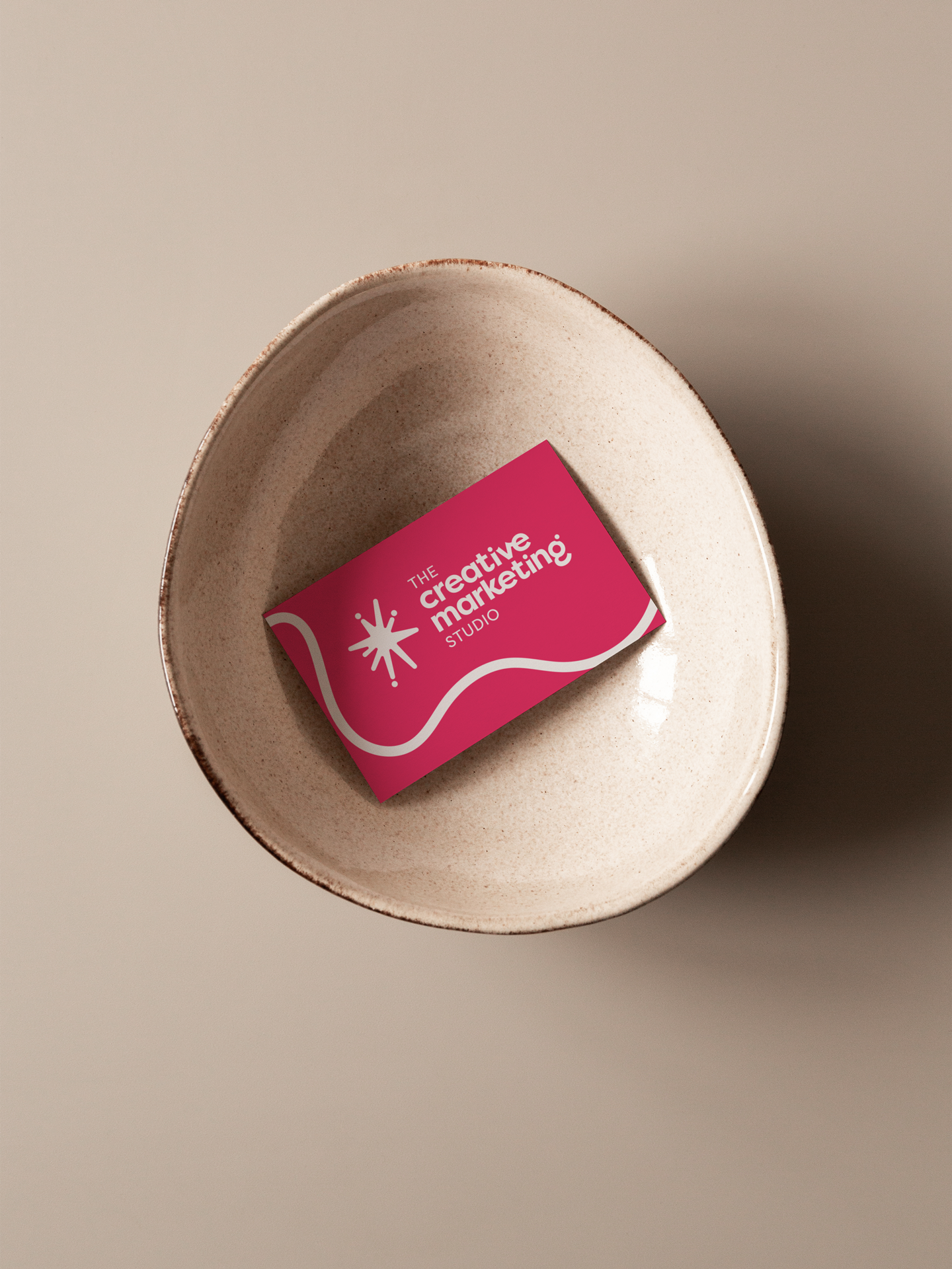 Close-up of a beige ceramic bowl with a pink business card inside that reads 'The Creative Marketing Studio' and features a white starburst logo.