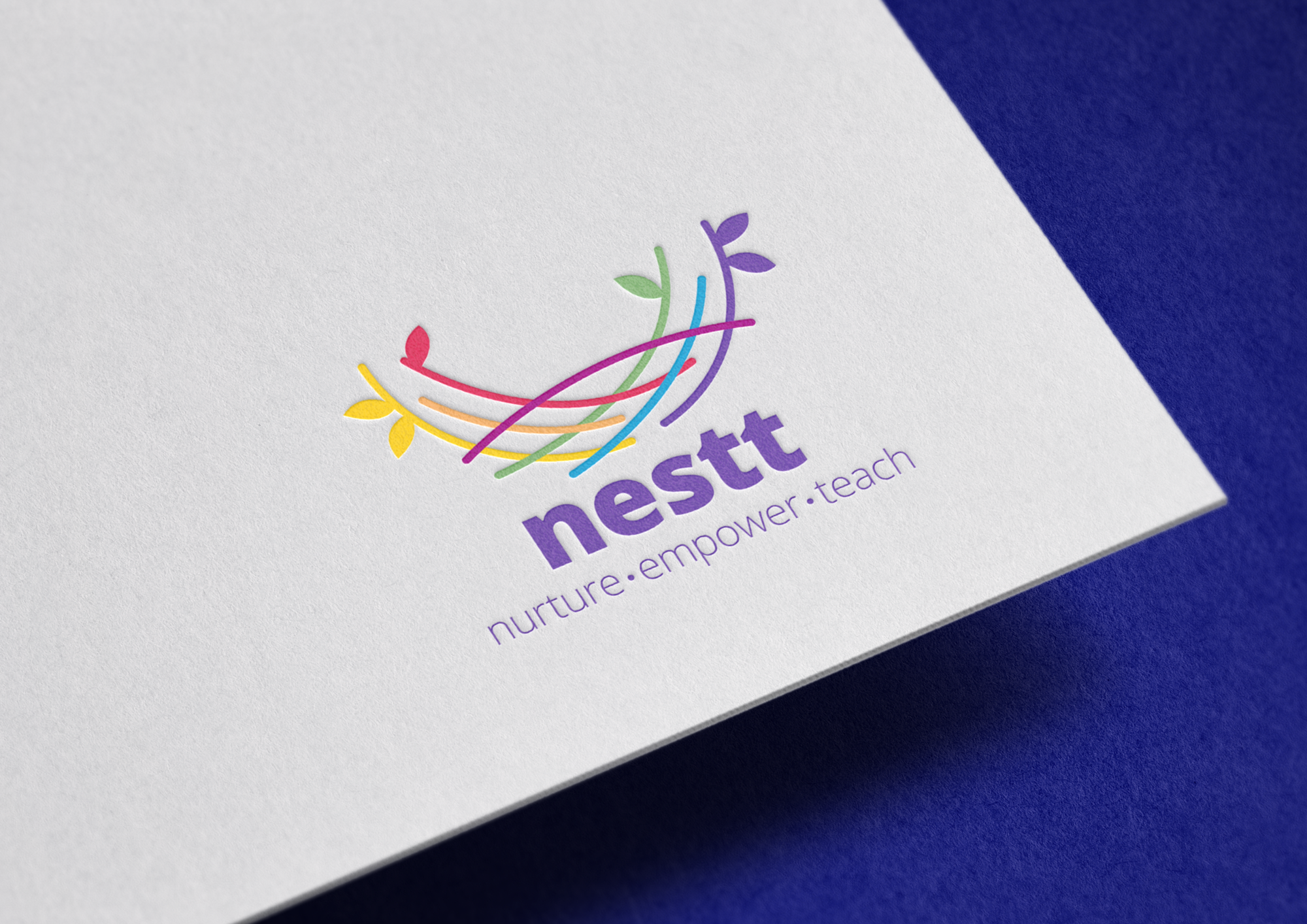 Close-up of a white card with a colorful logo featuring leaves and curved lines, and the text 'NESTT' and 'nurture • empower • teach' printed on it, placed on a dark blue background.