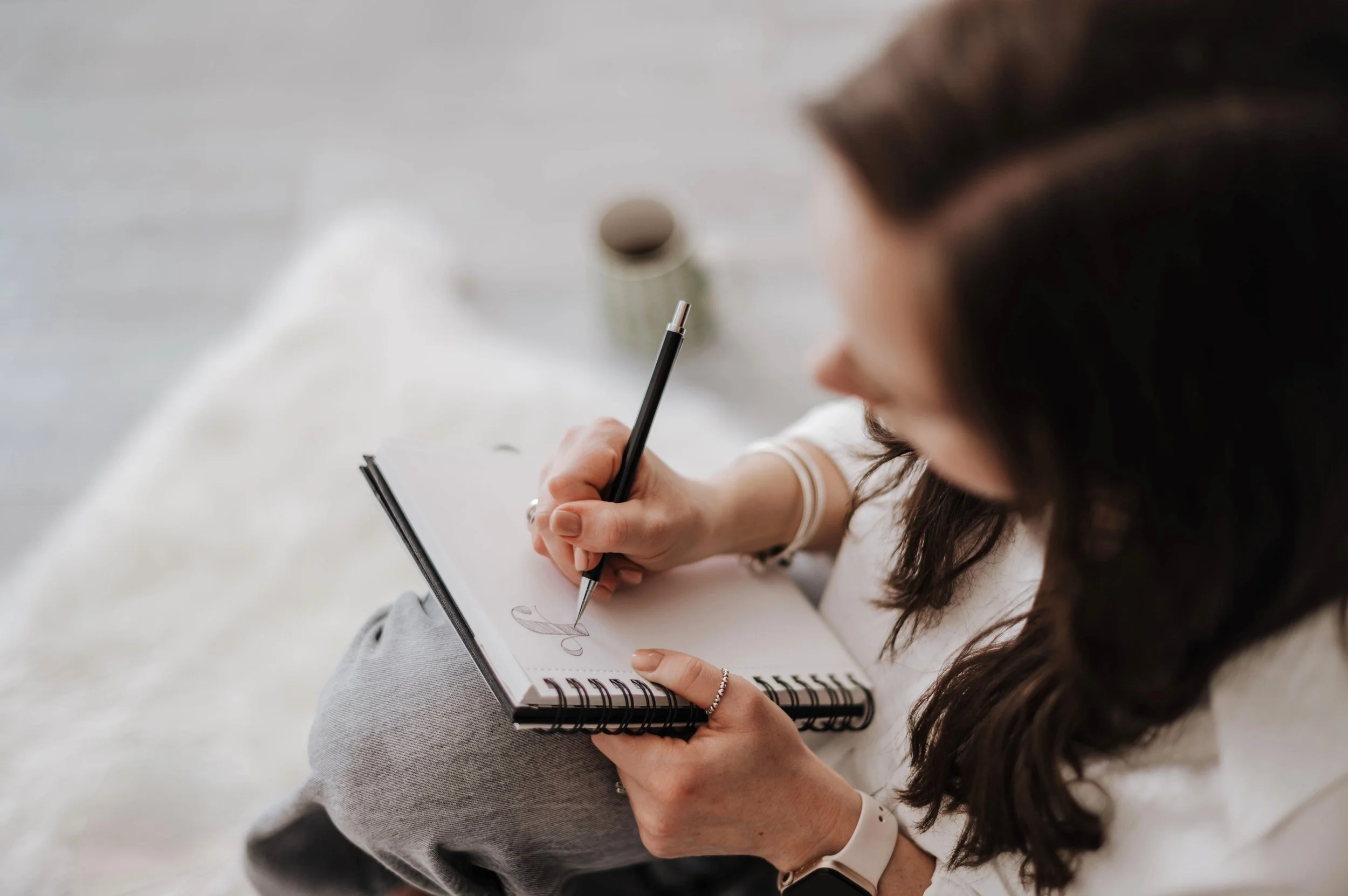 A brunette woman drawing an F letterform in a sketchbook with a black pencil, sitting cross-legged on the floor and focused on her artwork.