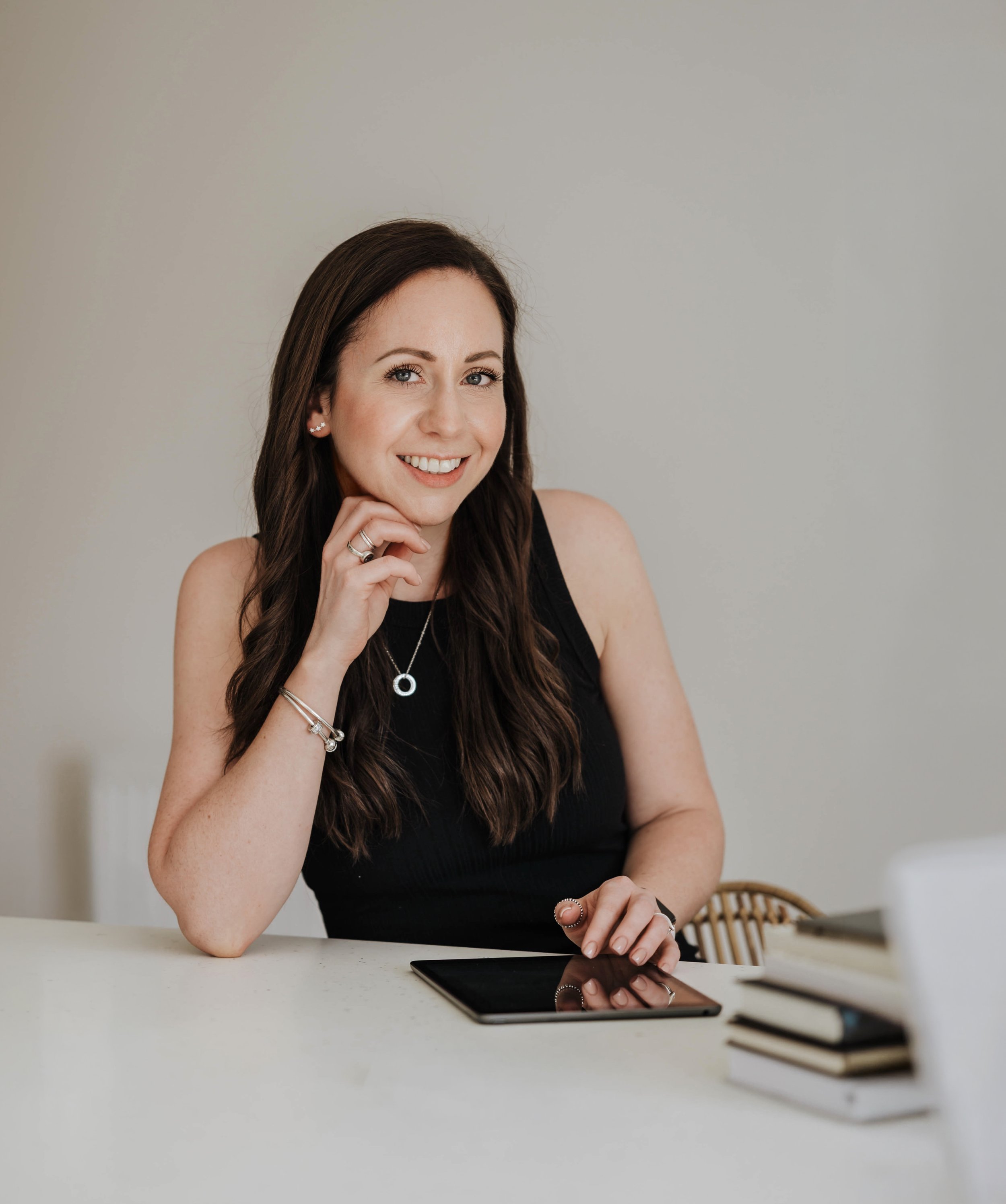 A young woman with long brown hair, smiling and resting her chin on her hand, sitting at a white table with a tablet and a stack of books, in a room with a neutral wall background.