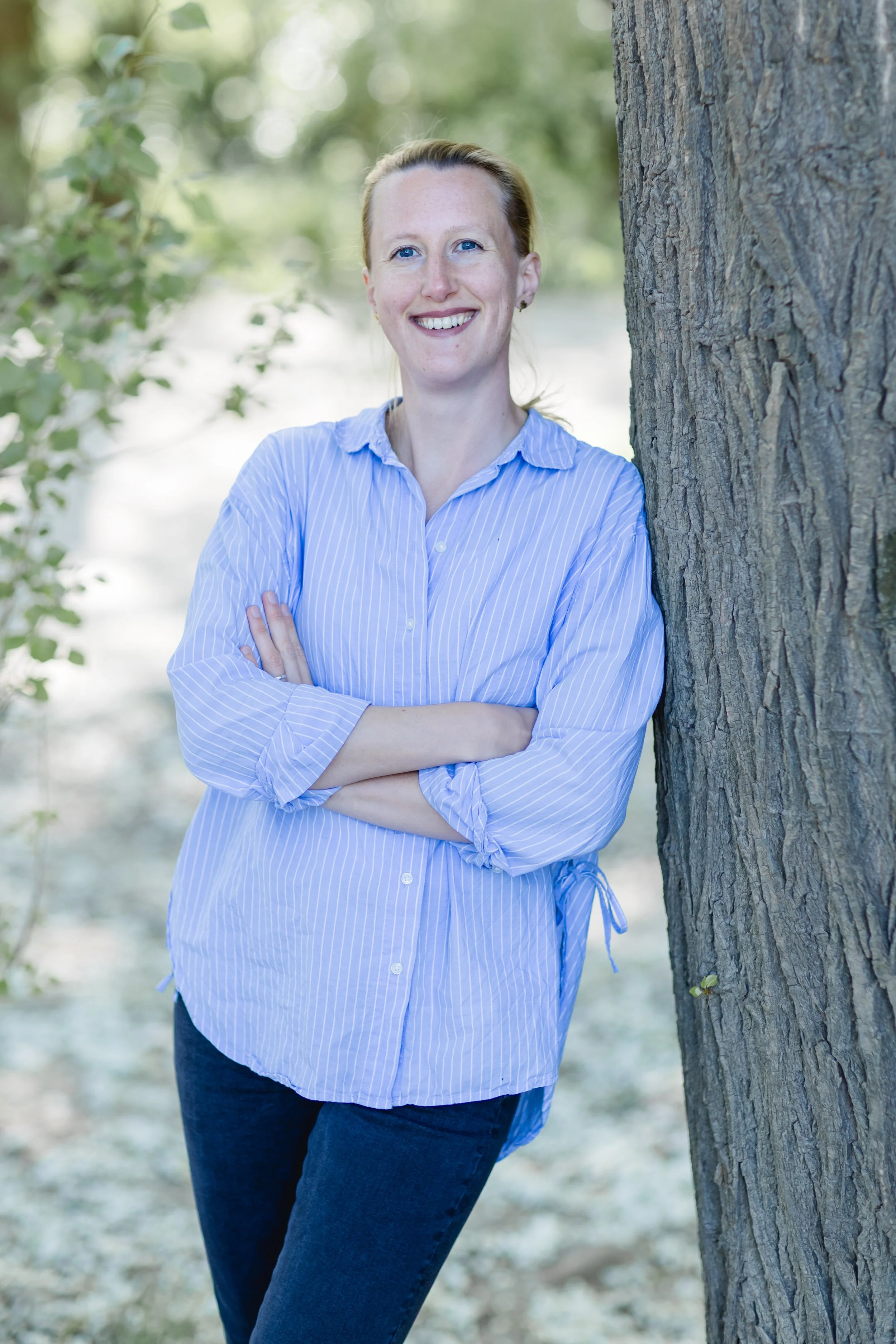 Eine Frau mit blonden Haaren, blauen Augen und einem blauen Streifenhemd lehnt an einem Baum im Park, lächelt und schaut in die Kamera.