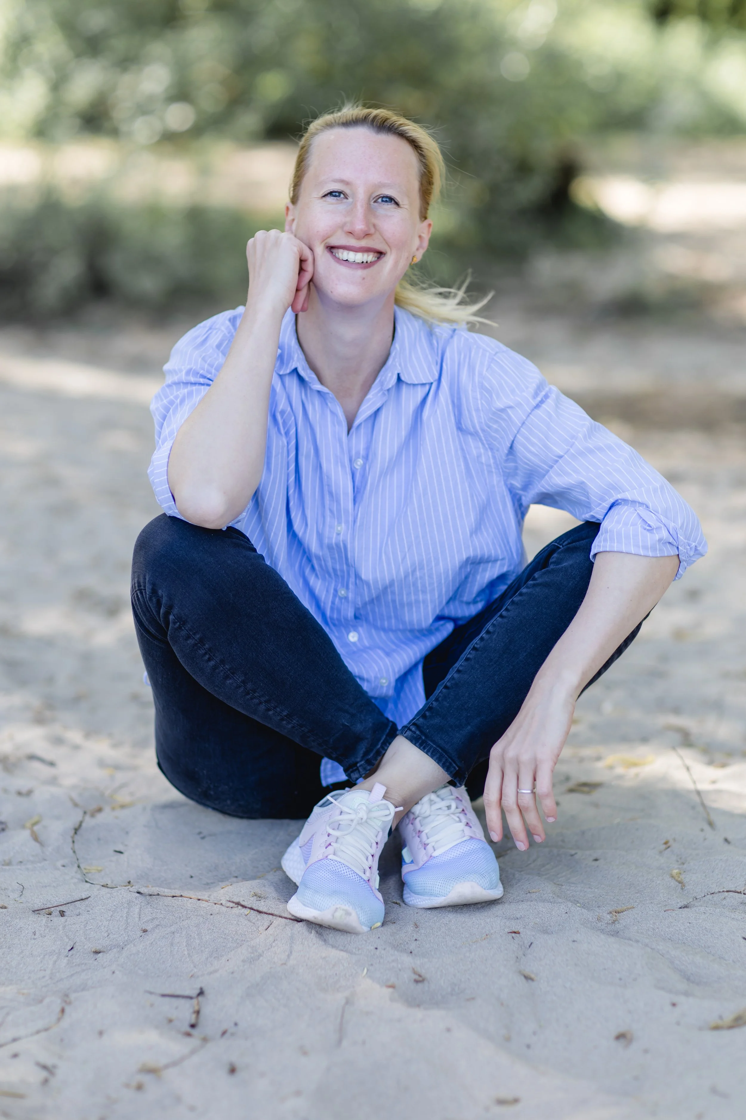 Lächelnde Frau mit blonden Haaren, blaue gestreifte Bluse, dunkle Jeans, weiße Turnschuhe, sitzt im Sand im Freien, hinter ihr ist grüne Vegetation, sonniger Tag.